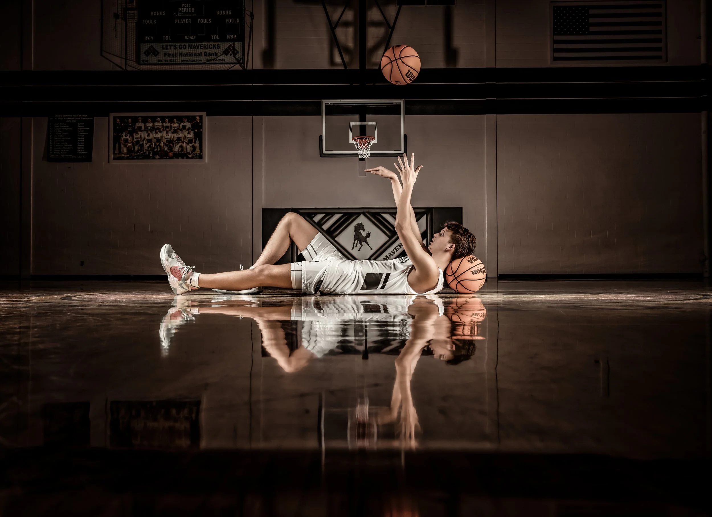A female basketball player lying on the court with basketballs around her, holding a basketball above her with one hand, inside a basketball gym.