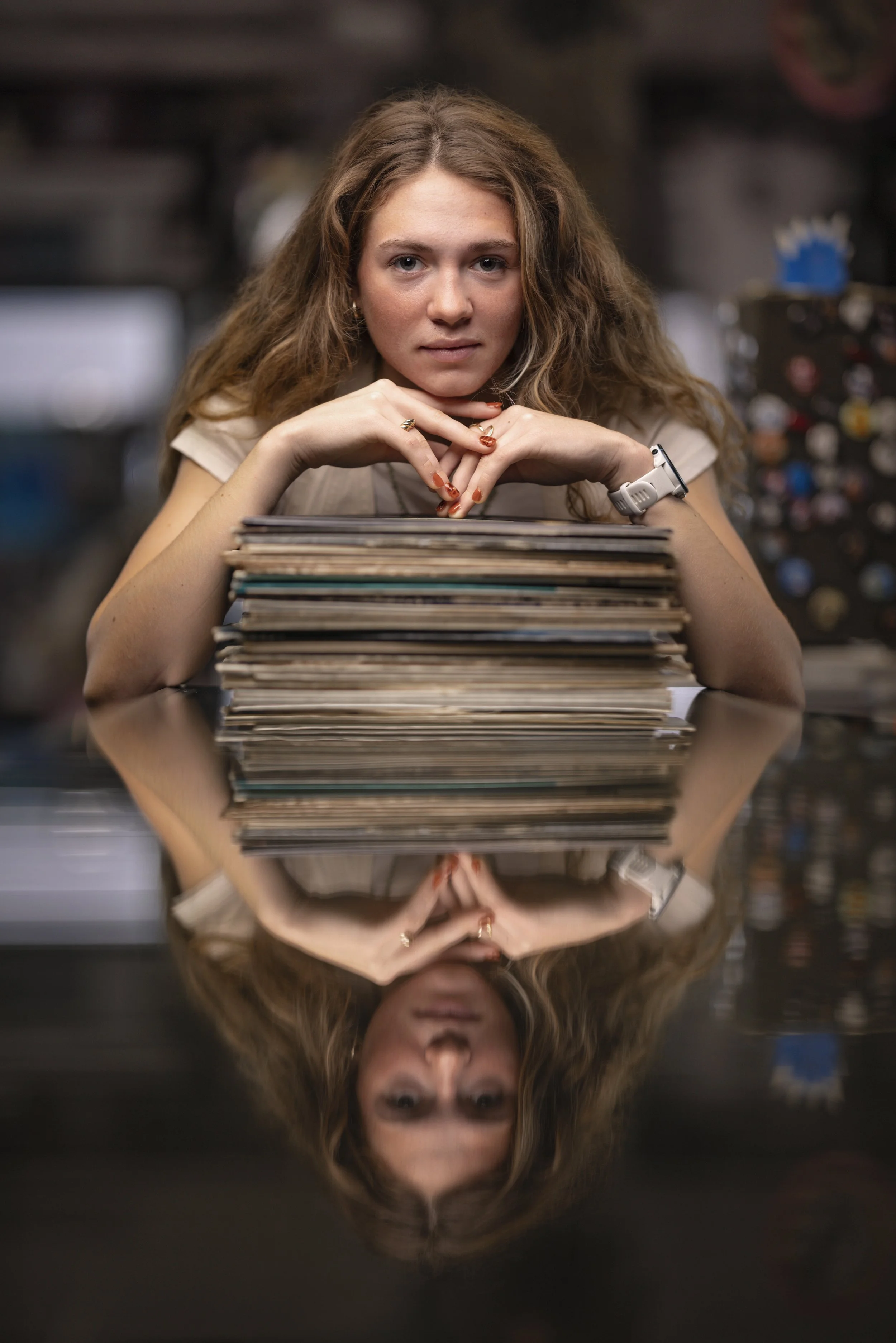 A young woman with long curly hair sitting at a table, resting her chin on her hands with fingers interlaced, looking directly at the camera. In front of her is a stack of vinyl records, and her reflection is visible on the shiny surface of the table