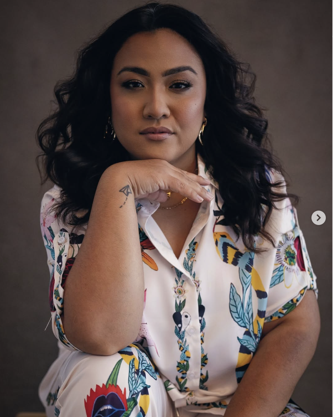 A woman with shoulder-length dark hair and medium skin tone, wearing a colorful floral blouse, is sitting with her chin resting on her hand, looking directly at the camera.