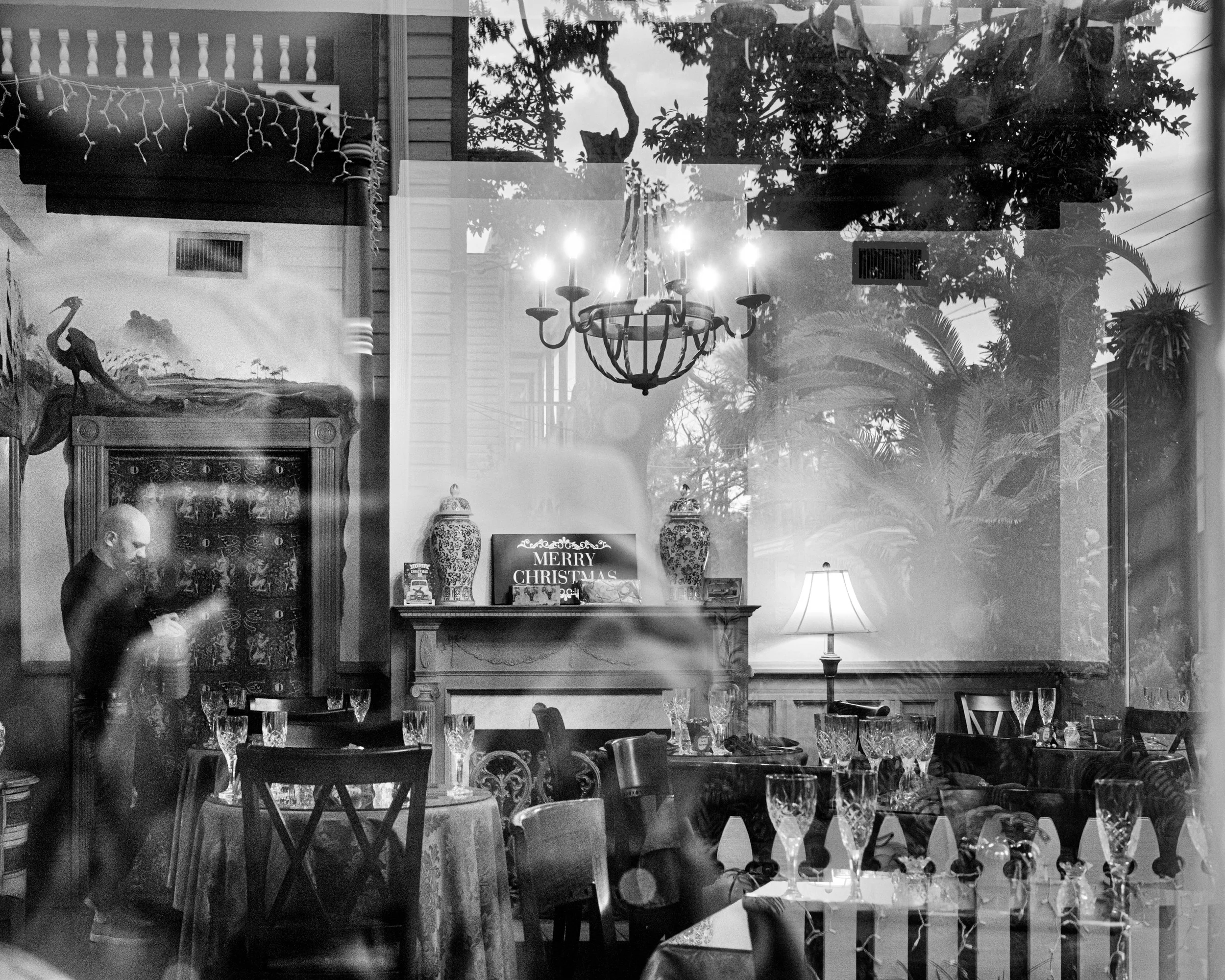 A decorated dining room seen through a window reflecting trees outside. The room has a chandelier, a lamp, a fireplace with a sign saying 'Merry Christmas,' and a table set with glassware and tablecloths. A person is standing near the table, possibly