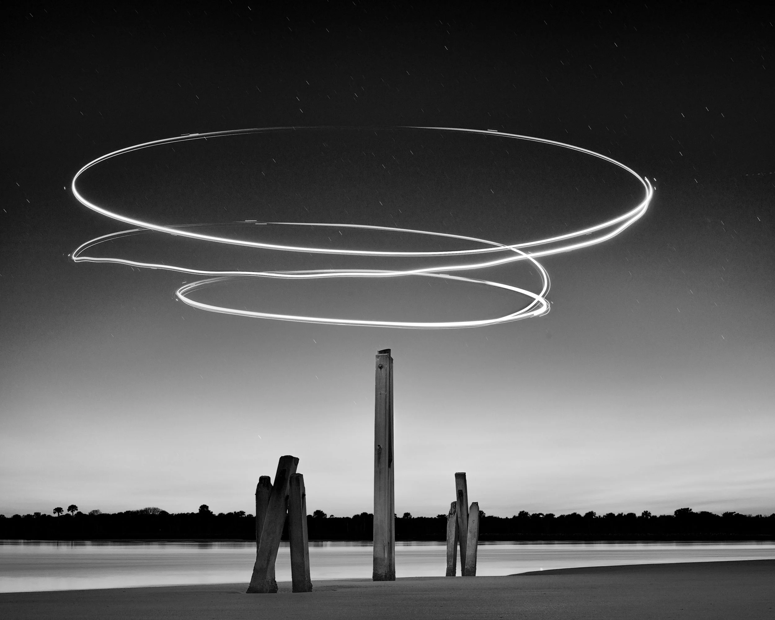 Long exposure photo of a night sky over a body of water with light trails produced by a circling drone, illuminated light streaks swirling above weathered wooden poles. Matanzas Inlet Palm Coast.