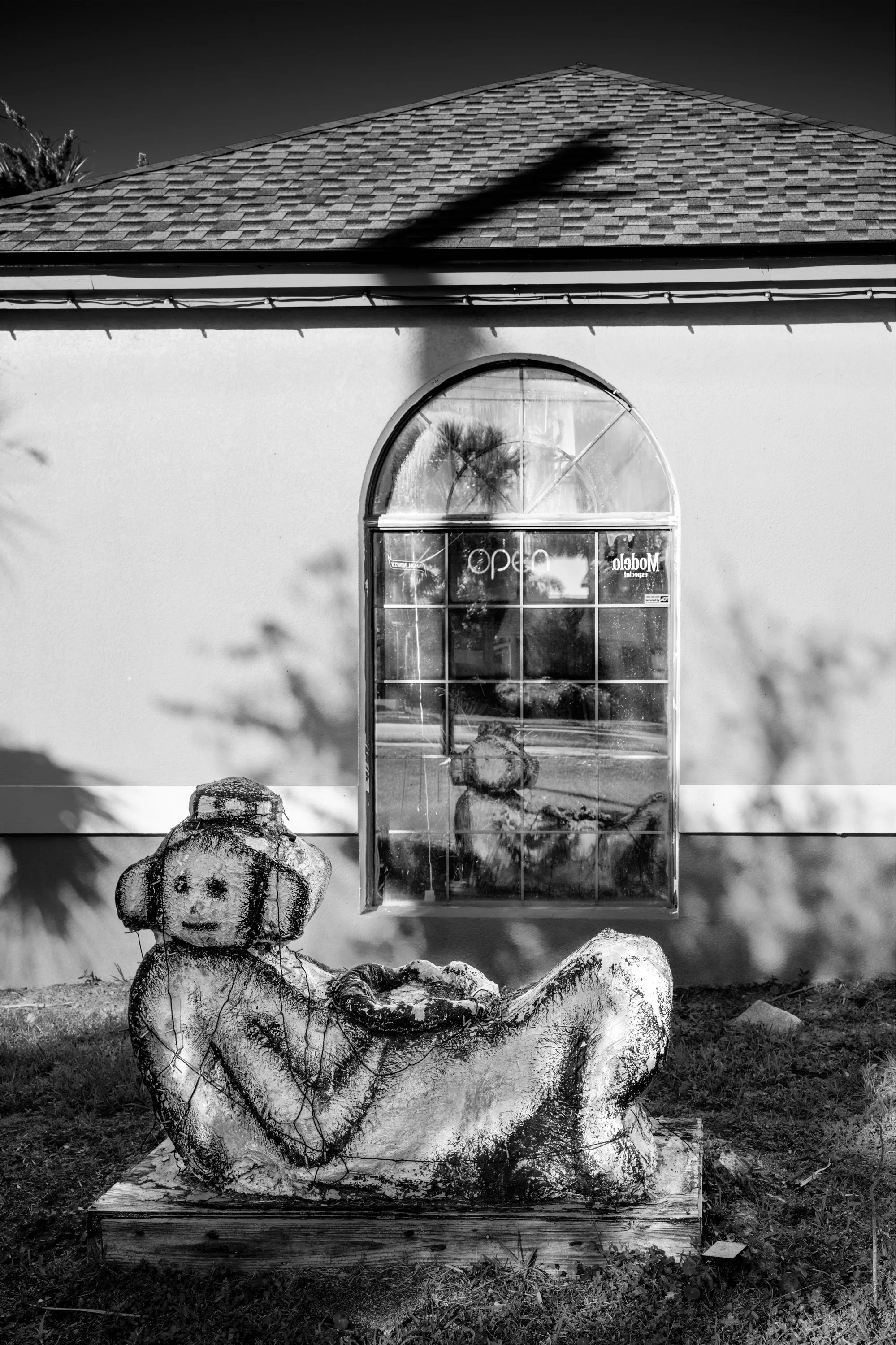 A black and white photo of a house with a shingled roof, a large arched window, and a sign that says "Open." In front of the house, there is a sculpture of a reclining dog made of a weathered material, possibly wood or concrete, placed on a wooden pl