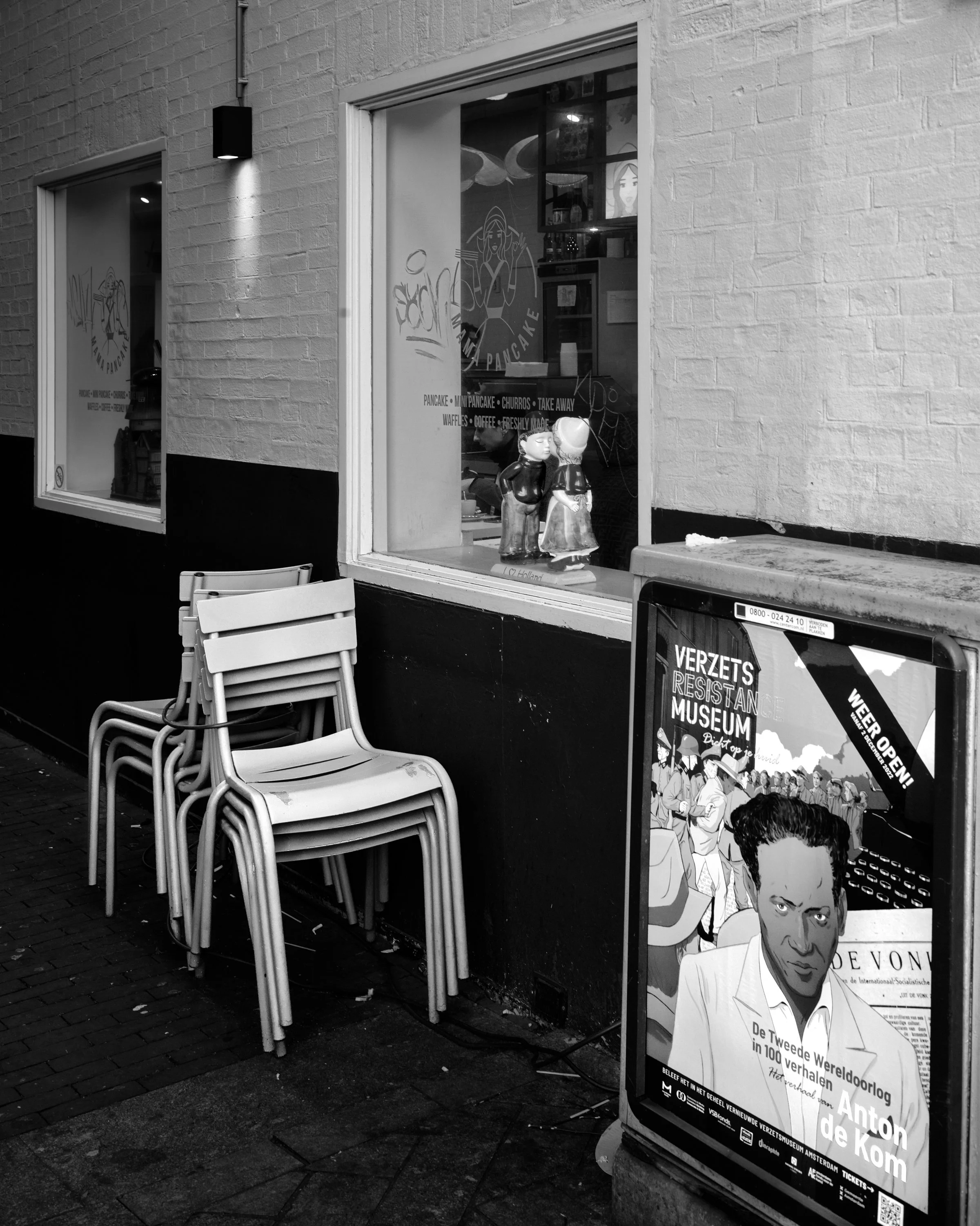 Black and white photo of a street scene from Amsterdam Netherlands with a stack of chairs near a window display showing two figurines, and a promotional poster for a resistance museum featuring a portrait of Anton de Kom. Hasselblad 
