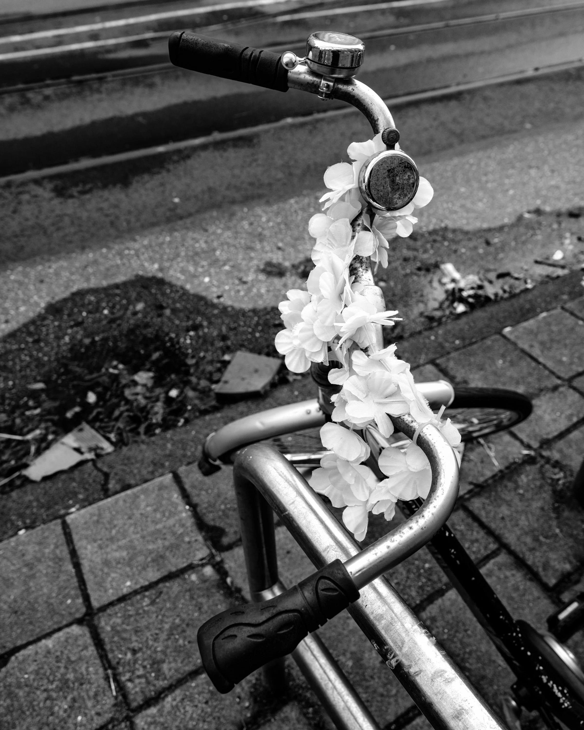 A bicycle decorated with a string of flowers leaning on a curb on a city sidewalk in black and white. Amsterdam
