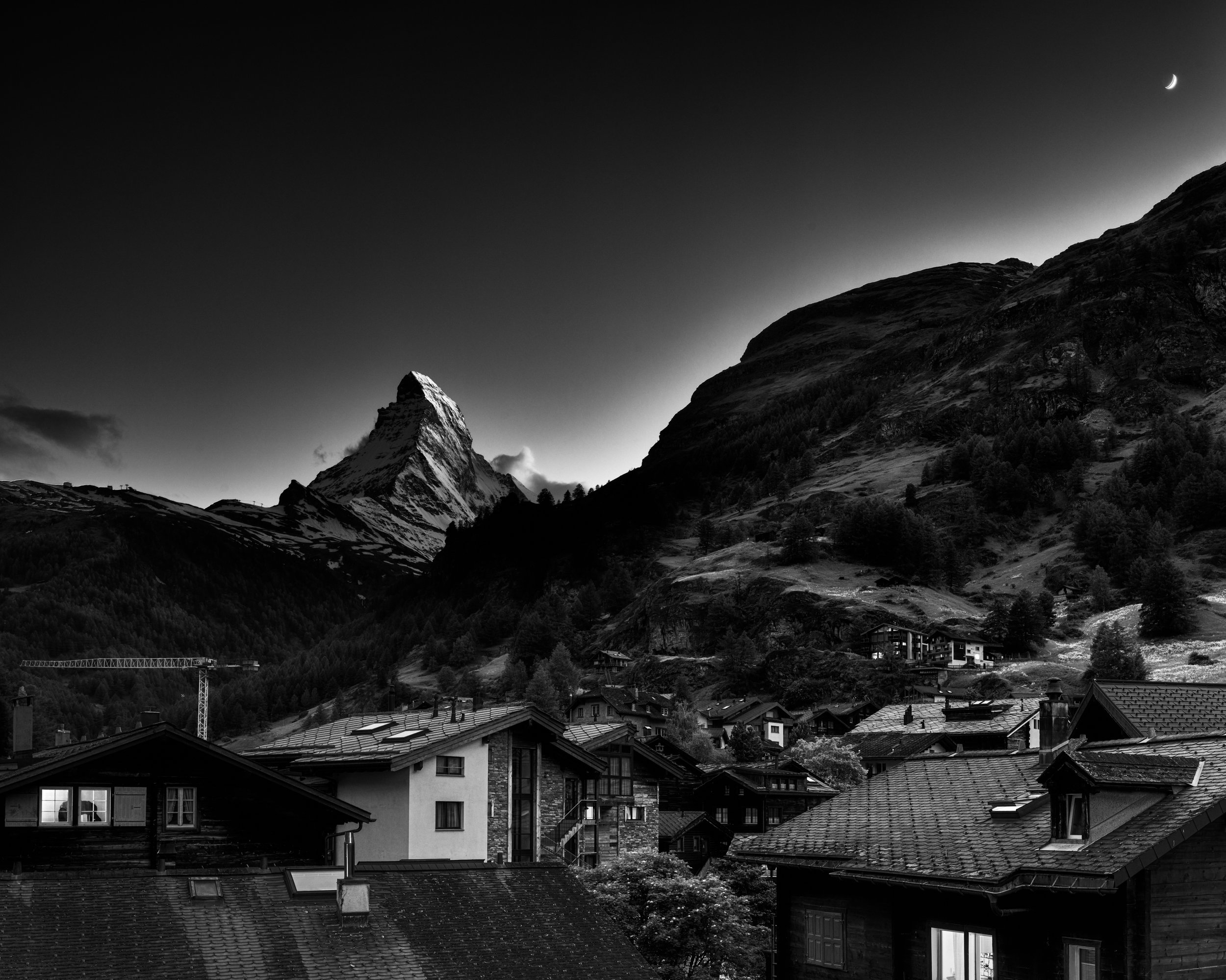 Black and white photo of Zermatt Switzerland with Matterhorn peak in the background, a crescent moon in the sky, and houses with tiled roofs in the foreground. Hasselblad CFV
