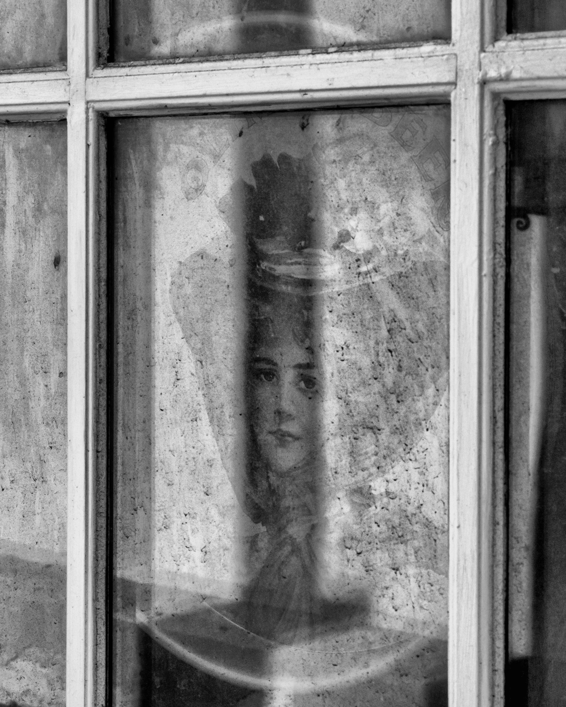 A black and white photo showing a pensive woman’s face visible surrounded by curtains through a dirty window. St Augustine