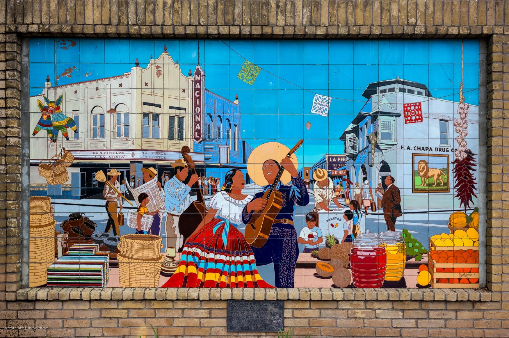 Colorful mural depicting a bustling town square with mariachi band, market stalls, piñata, and historic architecture.