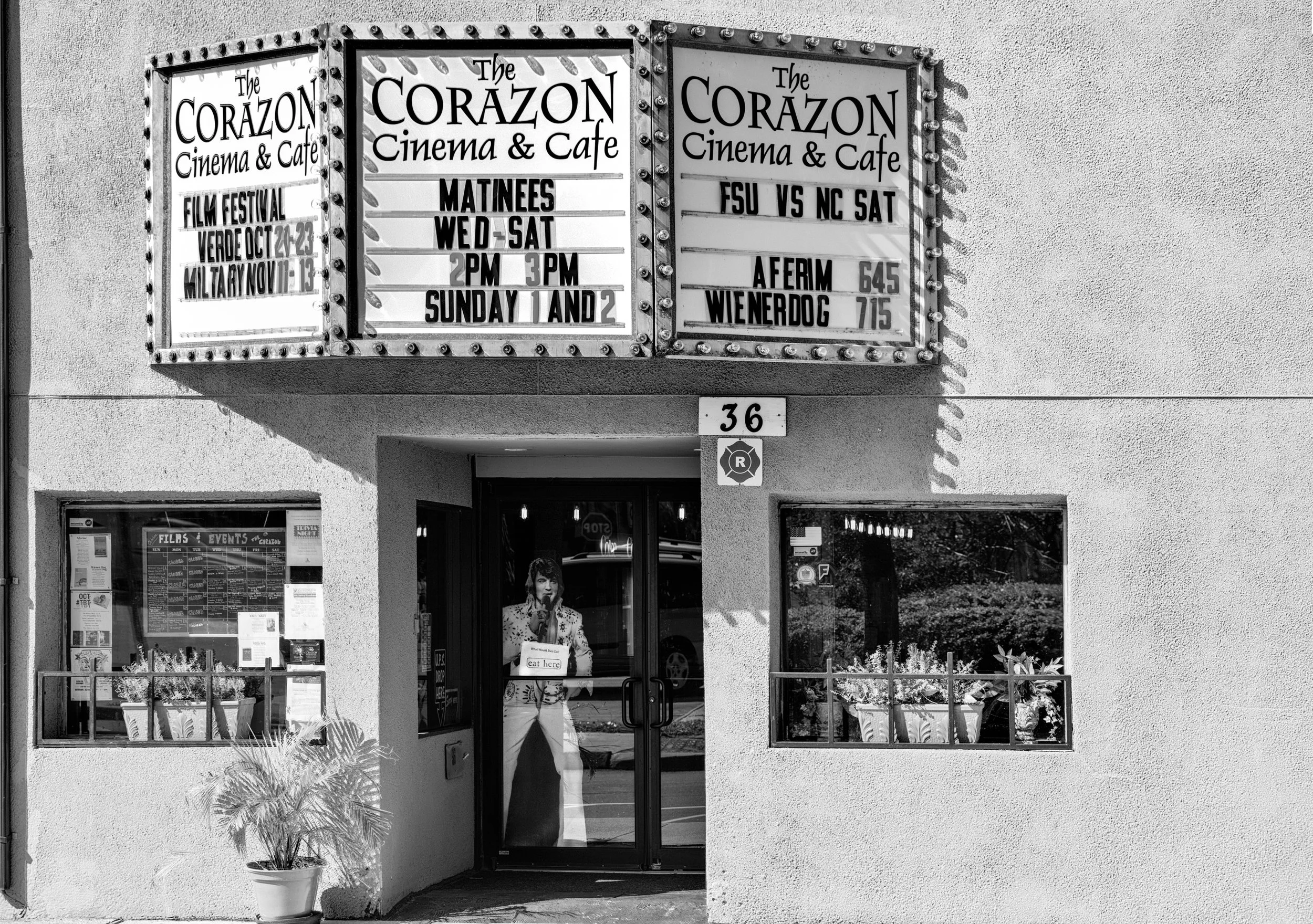 Black and white photo of the exterior of The Corazon Cinema & Cafe with marquee displaying film showtimes and events, Elvis Presley cutout at the entrance window in St Augustine Florida. This cinema was demolished after it shut down during COVID 19