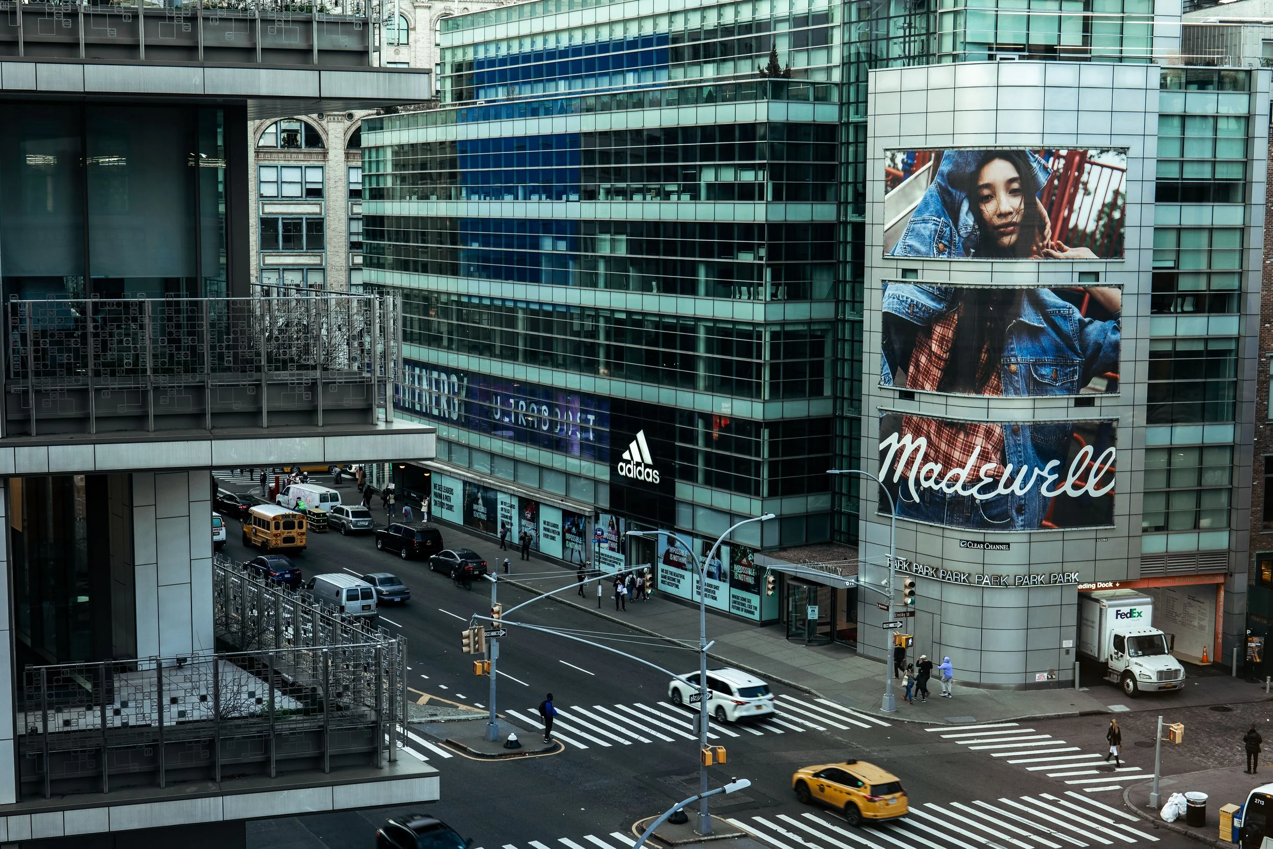 Urban street scene with Madewell and Adidas signage on buildings, traffic including a school bus and yellow cab, and a FedEx truck parked nearby.