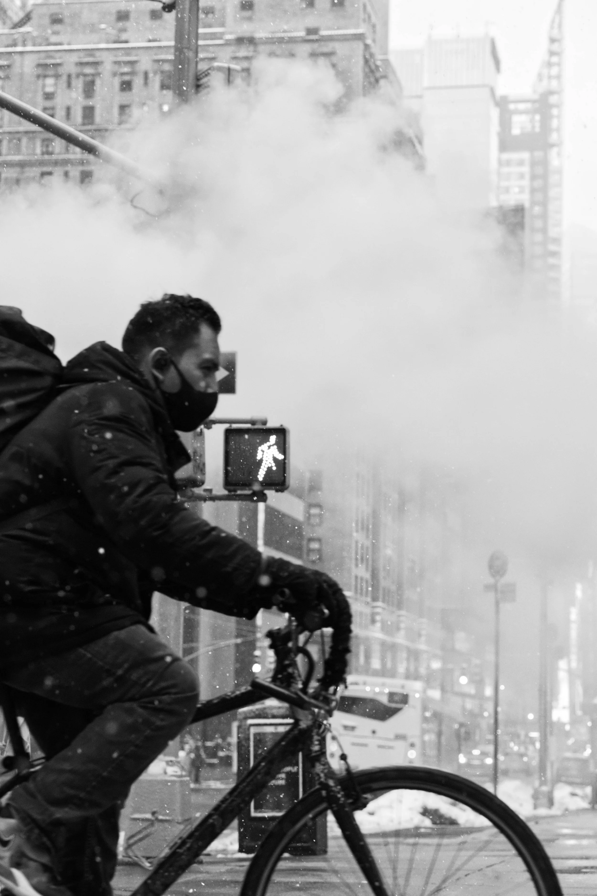 City street scene with a person on a bicycle wearing a mask, steam in the background, winter clothing, and a pedestrian crossing signal.
