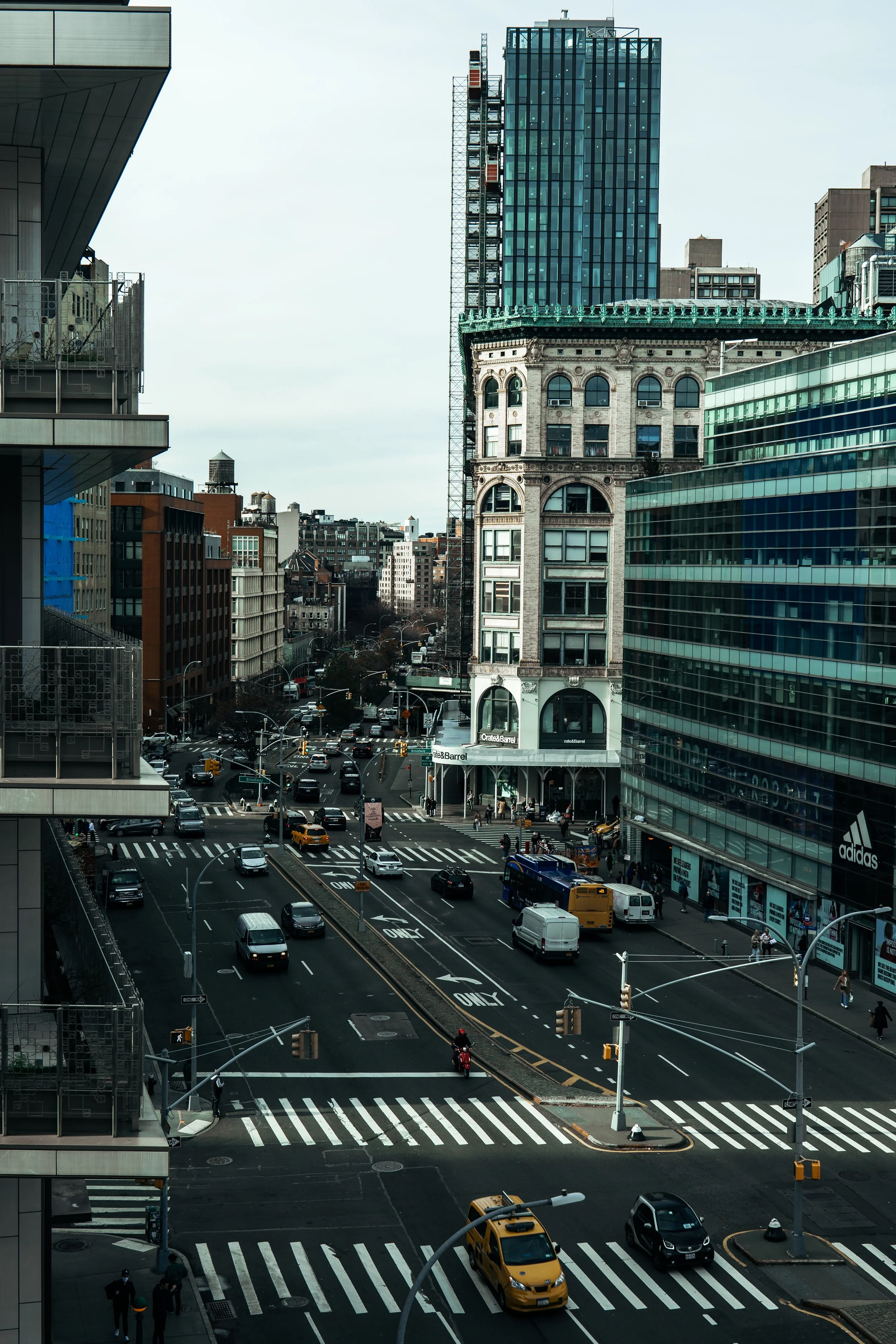 A busy urban intersection with multiple vehicles including yellow taxis, a motorcycle, and pedestrians. Surrounding buildings feature modern glass facades and traditional architecture. The scene is set in an urban environment with visible brand signa