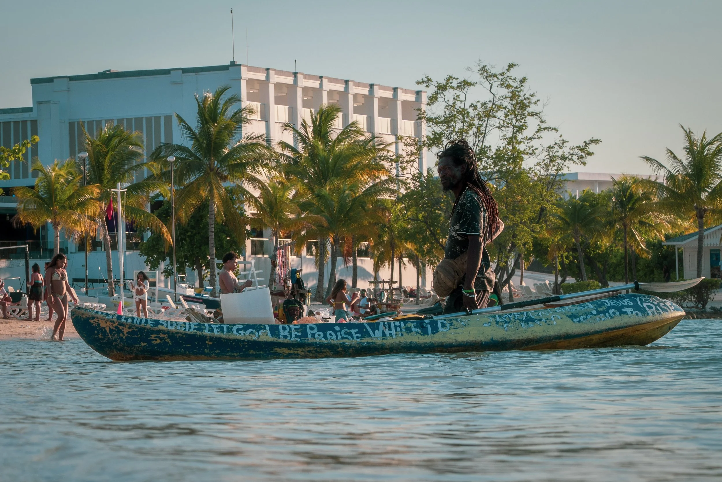 Man standing on a weathered boat in a tropical setting, with palm trees and a beach resort in the background, people sunbathing and swimming.