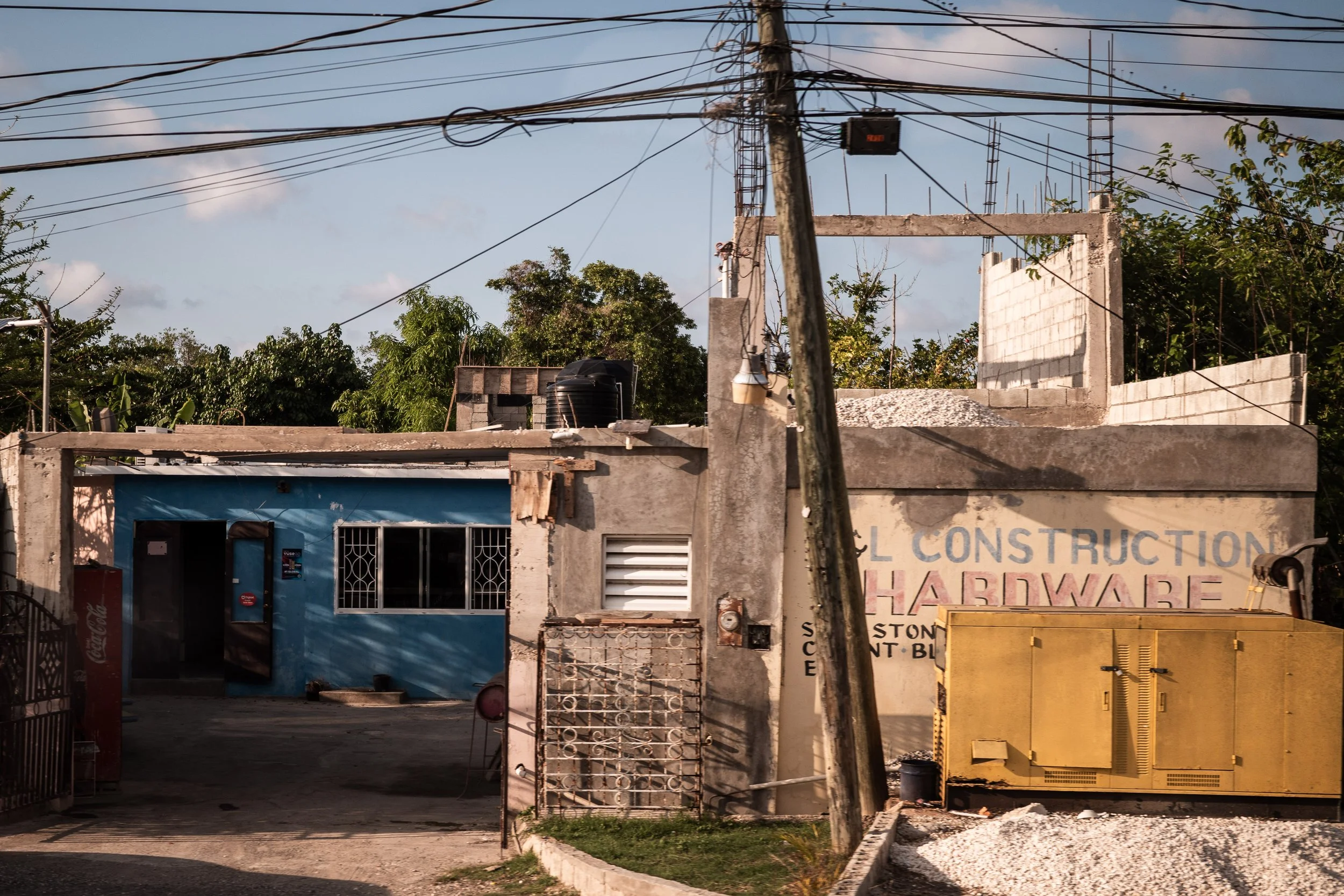 Outdoor scene showing a small cement building with a blue wall and a glass window, electric wires overhead, and a faded sign reading 'Hardware.' Trees and a clear sky in the background.