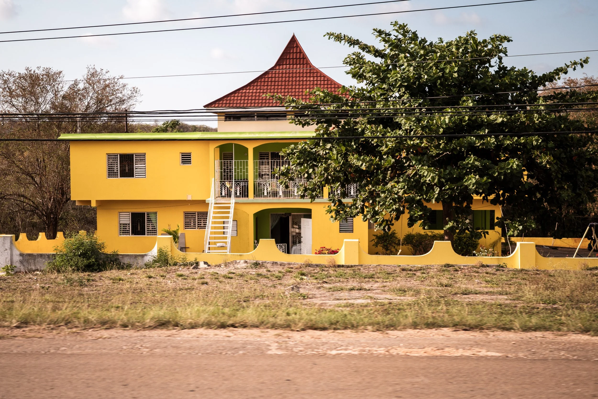 Yellow two-story building with green accents, a red roof, and white stairs in front, surrounded by a low wall and trees.