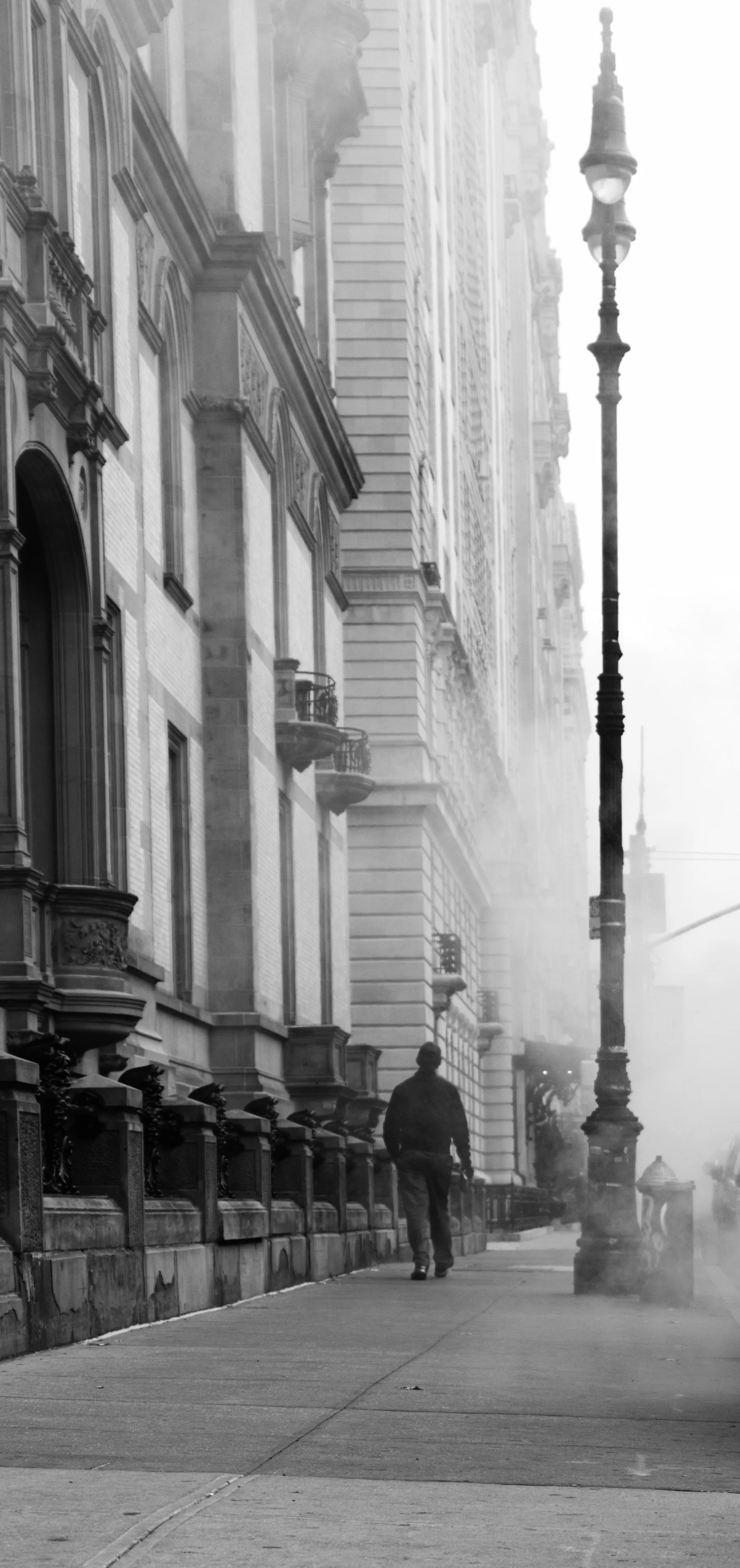 Black and white photo of a person walking down a city sidewalk next to tall buildings, with steam rising and a vintage street lamp.