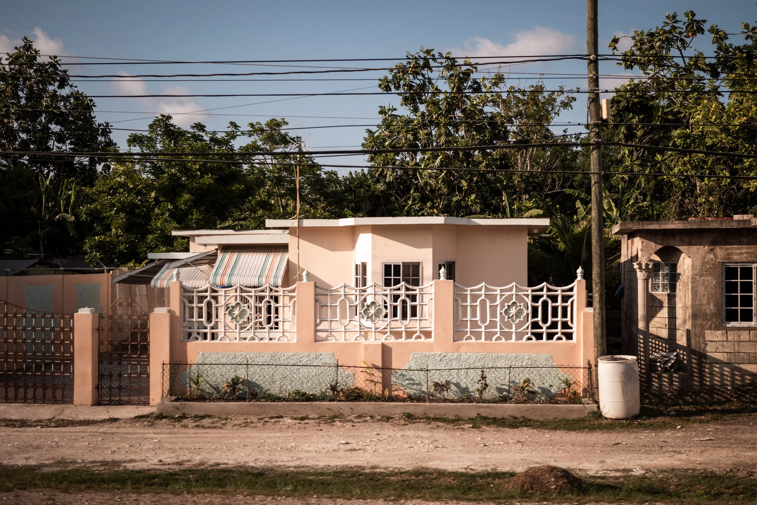 A small, light-colored house with decorative wrought iron fencing and a striped awning, situated in a residential area with trees and power lines.