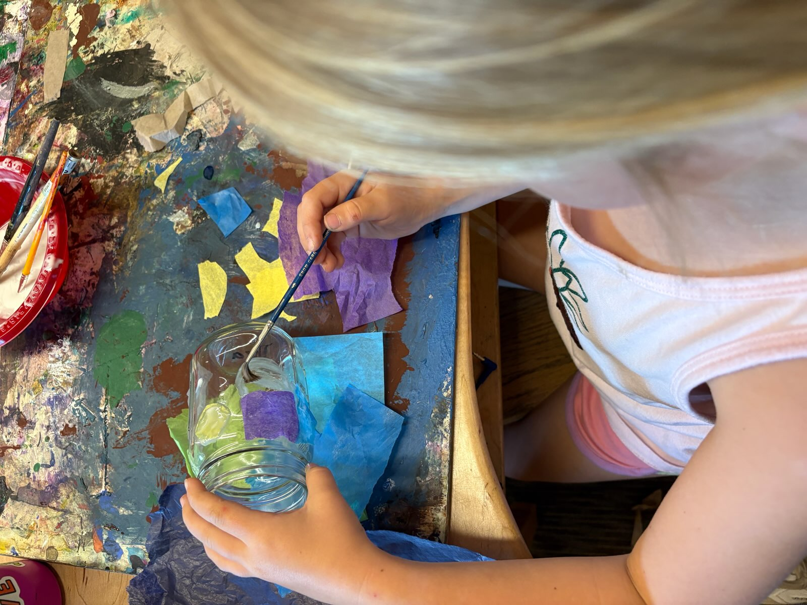 Birds eye view of a child applying tissue paper to a jar with a paintbrush to create a lantern
