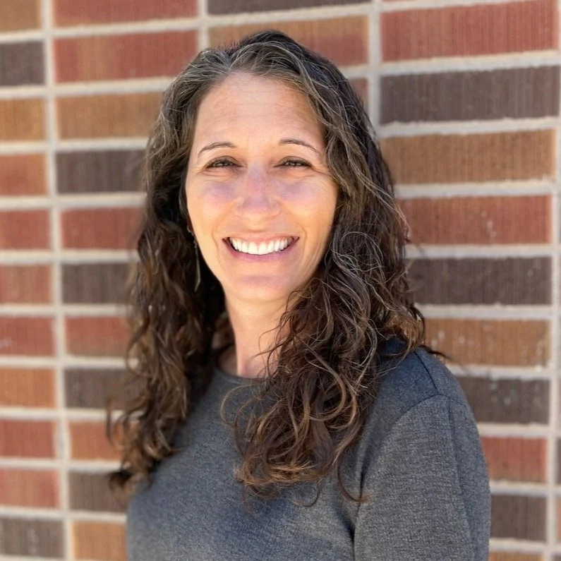 A smiling woman with long, curly brown hair standing in front of a brick wall, wearing a dark gray top.