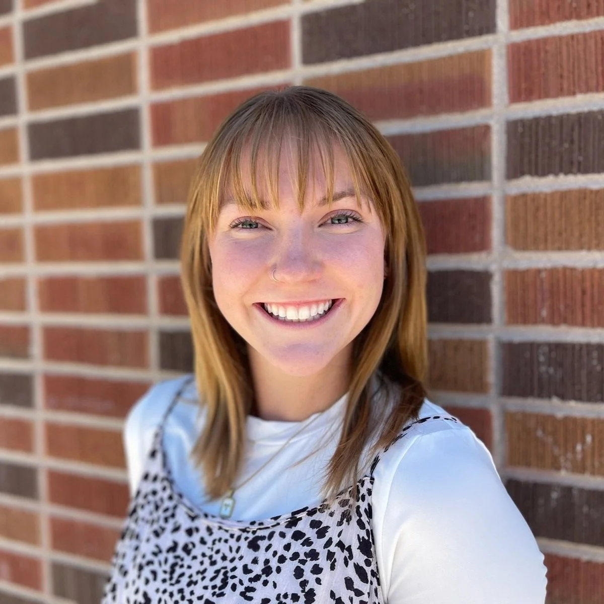 A young woman with reddish-brown hair, wearing a white top and a leopard print dress, smiling in front of a brick wall.