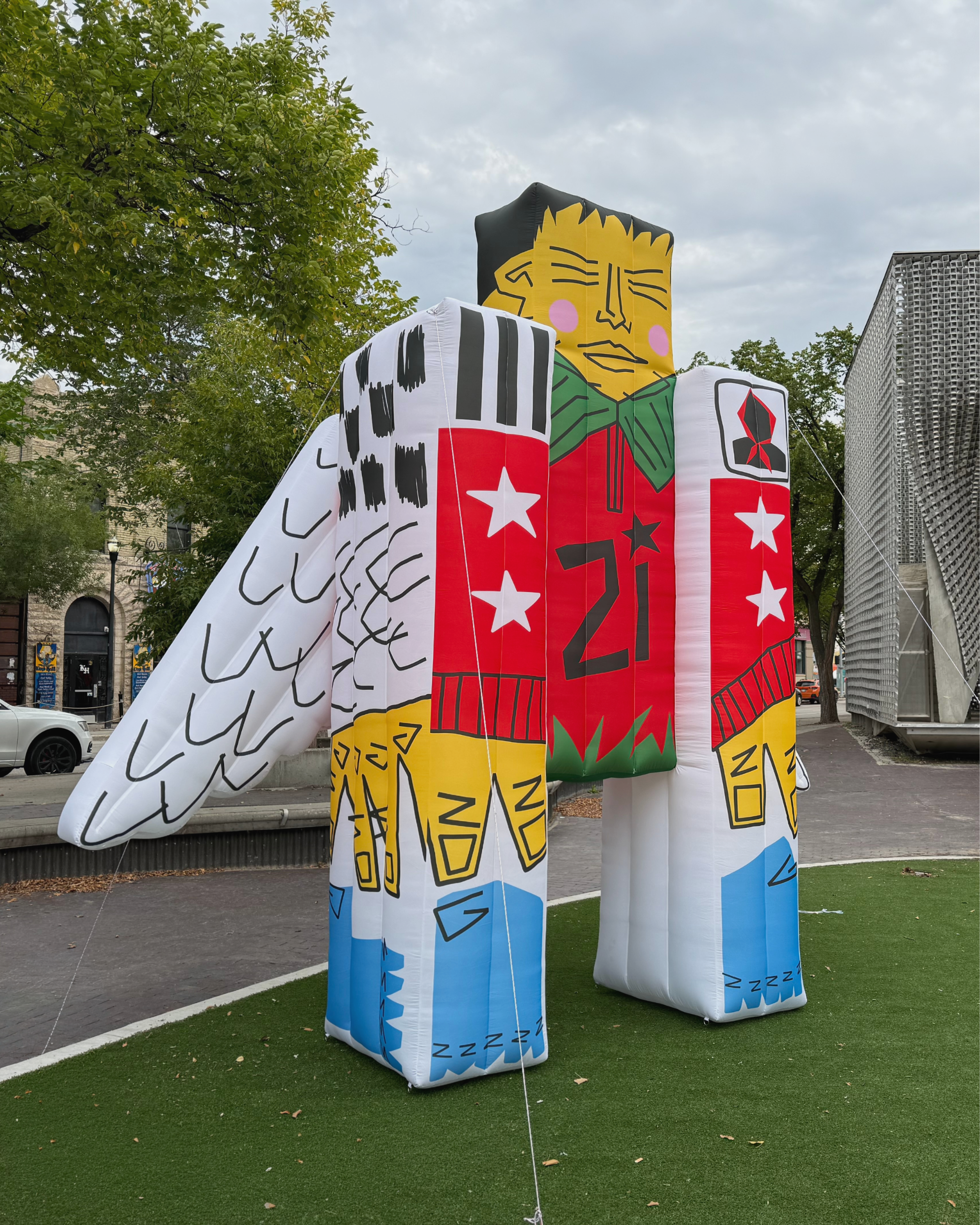 Colorful large art installation resembling a human figure, with geometric and abstract markings, situated outdoors on a grassy area next to a sidewalk and street, with trees and buildings in the background.