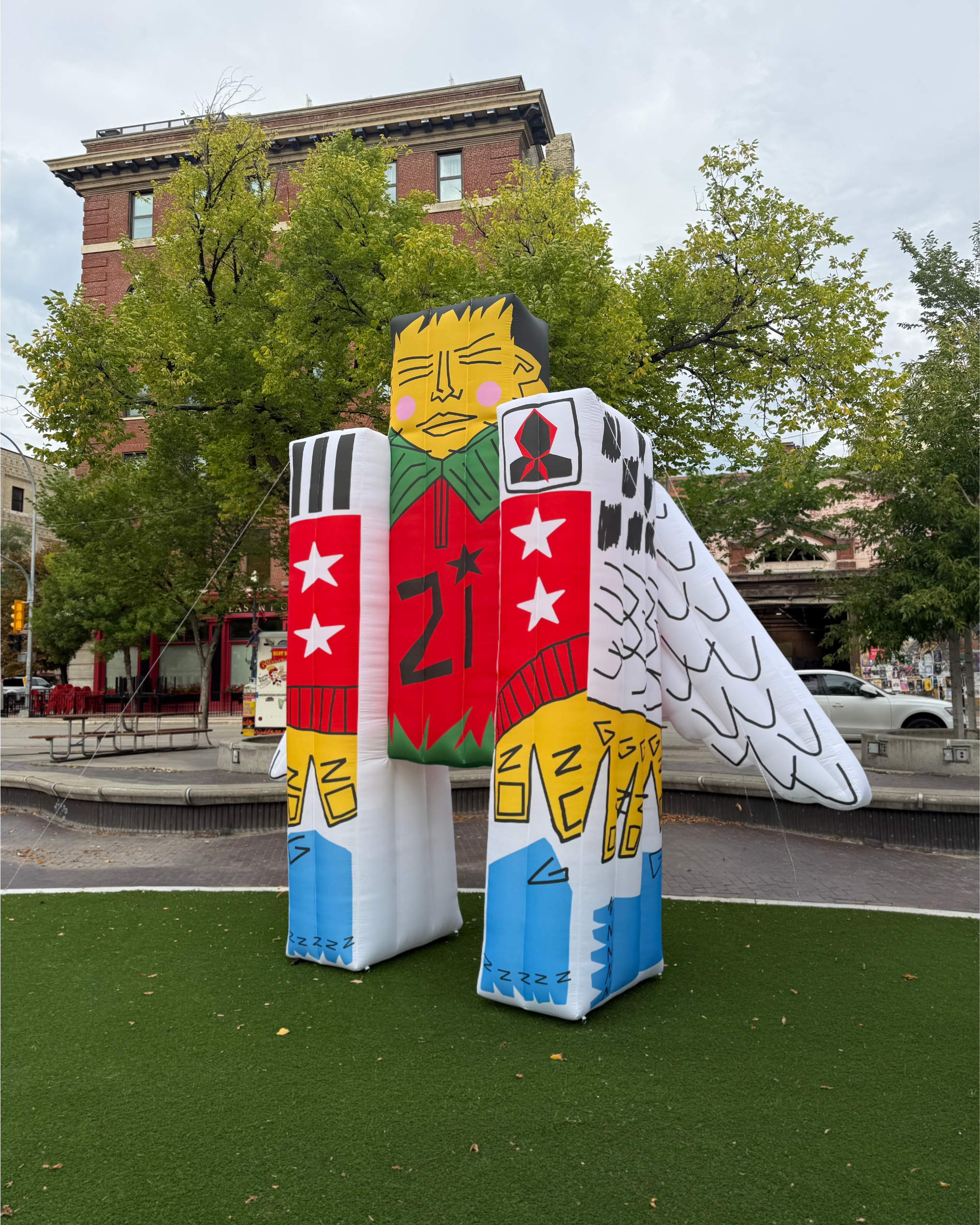Colorful inflatable structures designed to resemble a person with a yellow face, black hair, and pink cheeks, decorated with various patterns and symbols, set up outdoors on a grassy area in an urban environment.