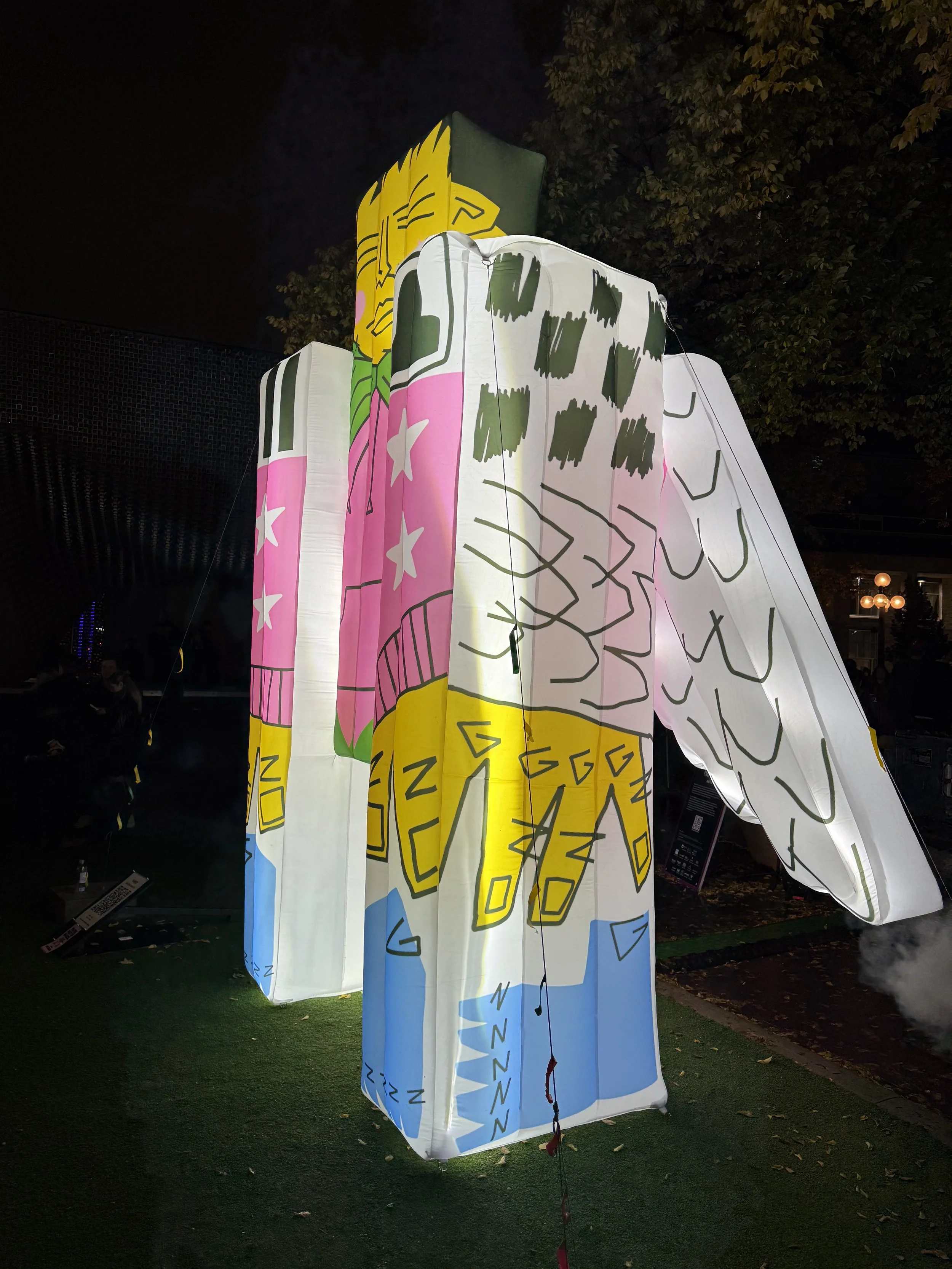 Illuminated large paper lanterns with colorful abstract and cartoonish designs are displayed outdoors at night, surrounded by trees and a tiled structure in the background.