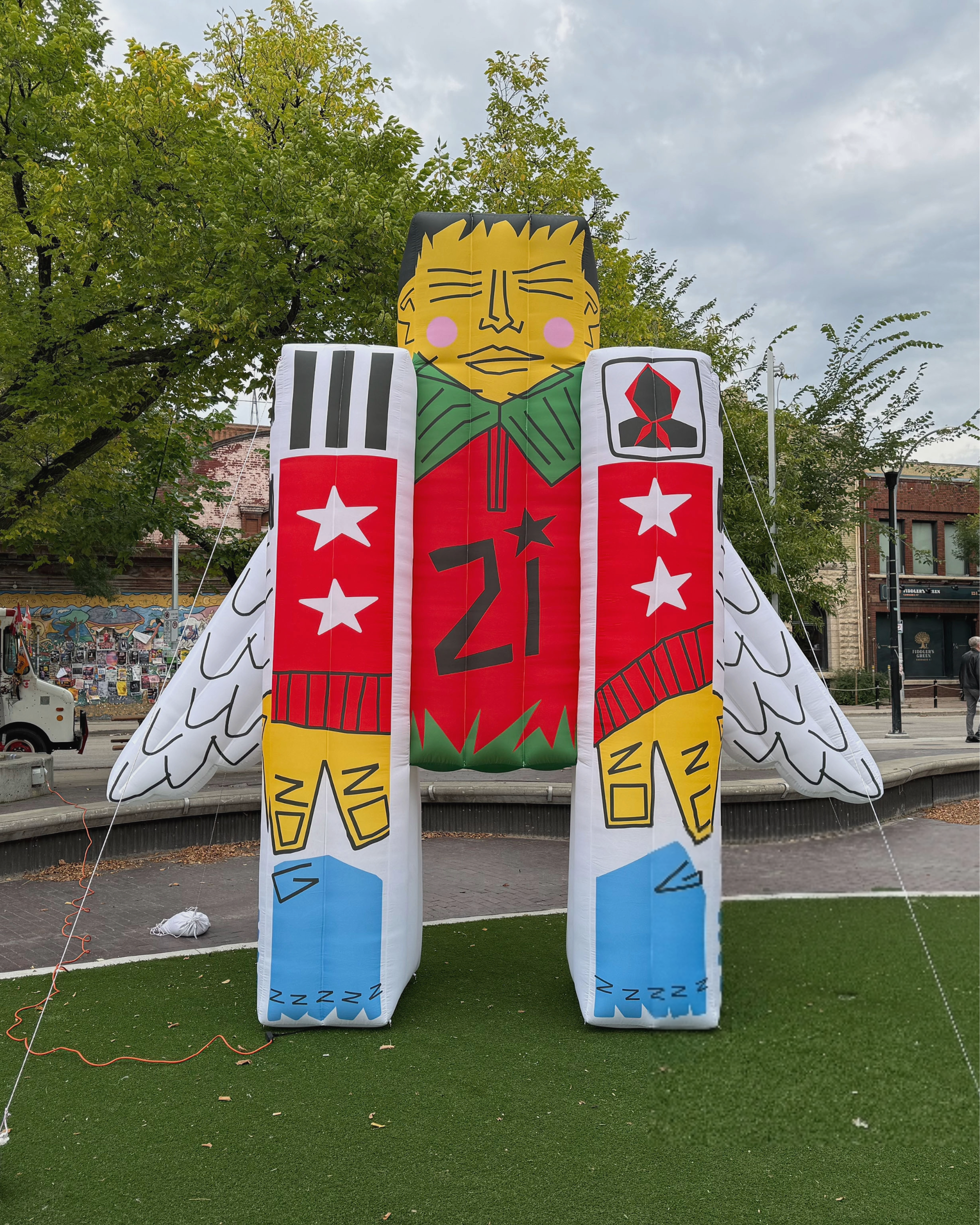 Colorful inflatable sculpture of a person with wings, with a large face at the top, standing on a patch of grass in an outdoor setting, with trees and buildings in the background.