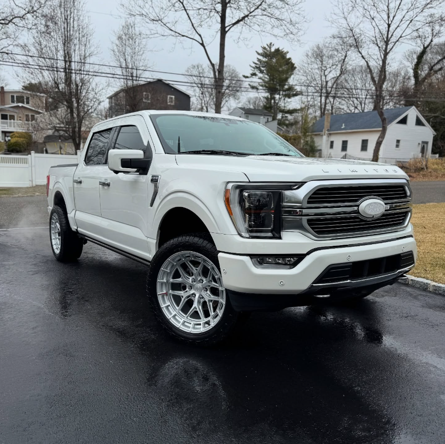 White Ford F-150 Limited crew cab pickup truck with chrome grille, lifted suspension, and polished aftermarket wheels on a wet driveway