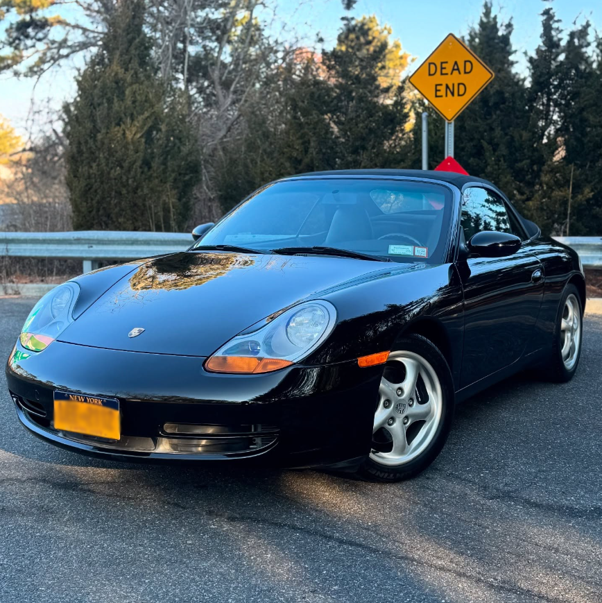 Black Porsche Boxster 986 roadster with soft top, round headlights, and silver five-spoke alloy wheels parked outdoors