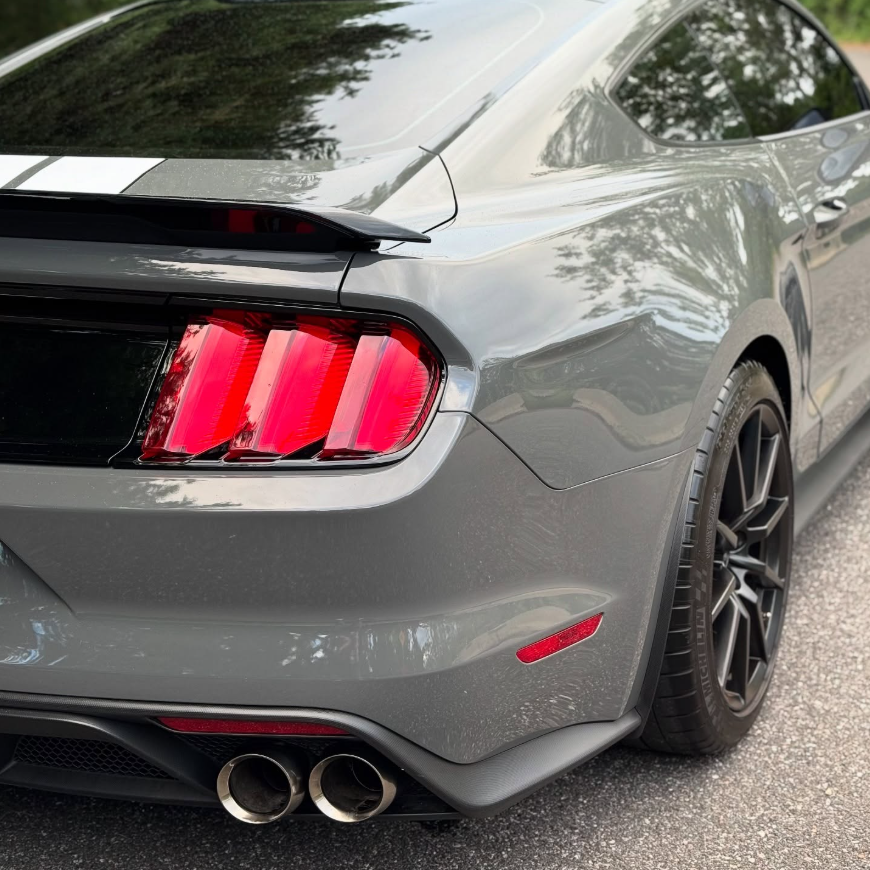 Rear quarter view of a gray Ford Shelby GT350 Mustang showing tri-bar taillights, rear spoiler, quad exhaust tips, and black wheels