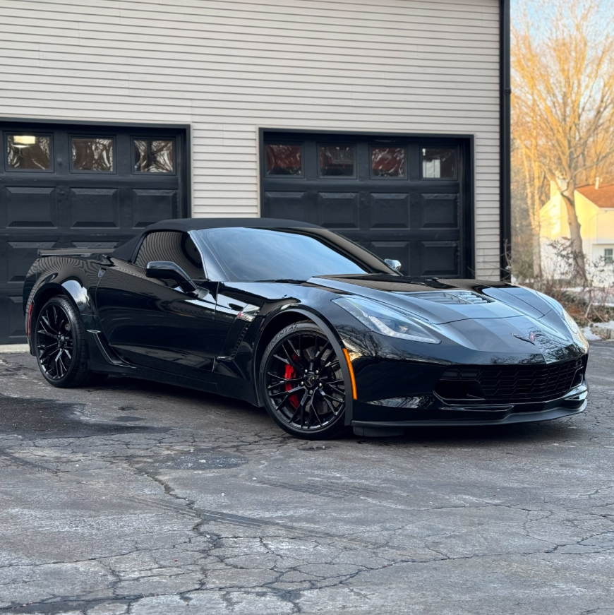 Black Chevrolet Corvette C7 Z06 convertible with soft top, front splitter, red brake calipers, and gloss black wheels parked in front of a garage