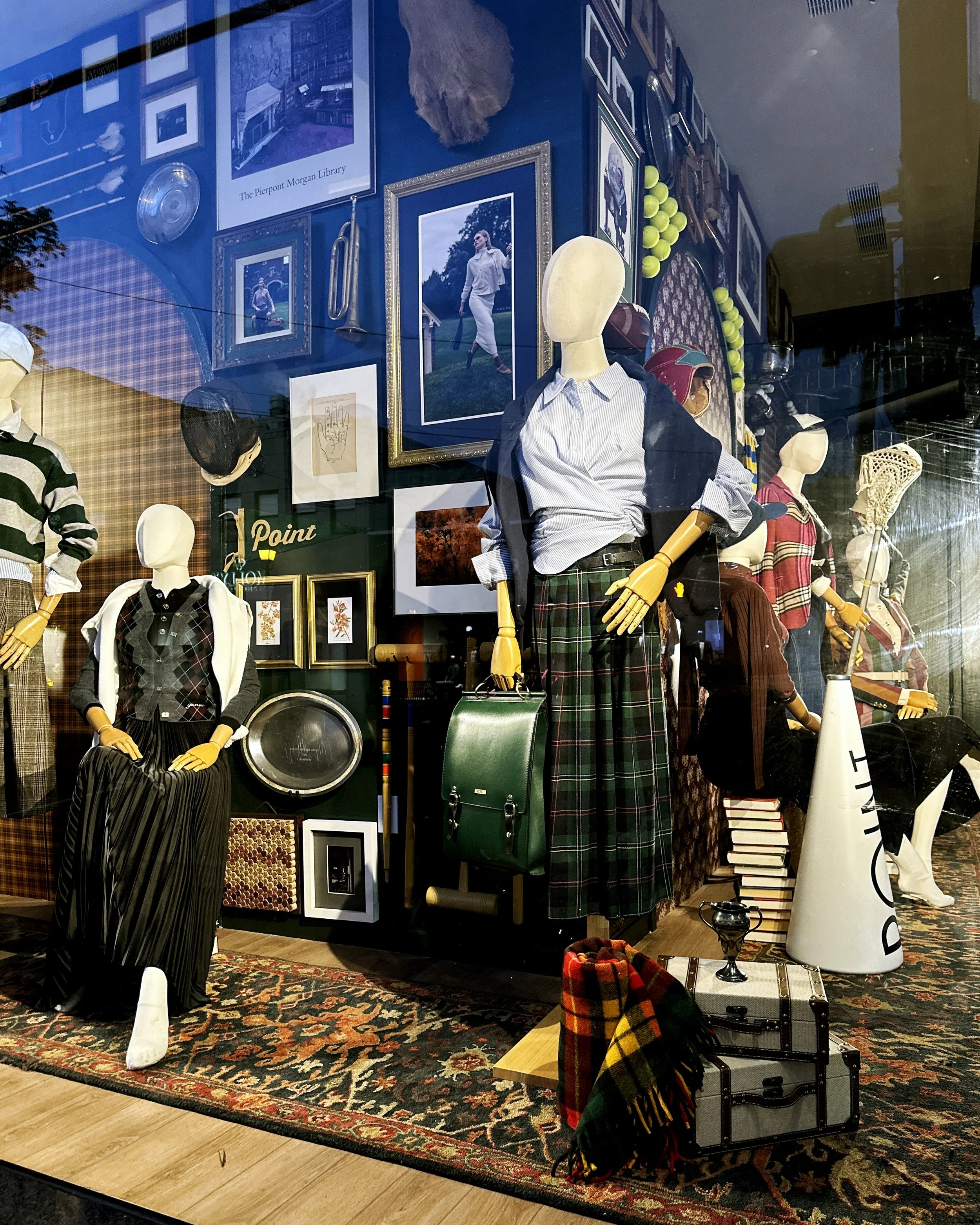 A corner retail window display with a preppy, collegiate theme, featuring mannequins in classic plaid and striped clothing, vintage props, and framed art.