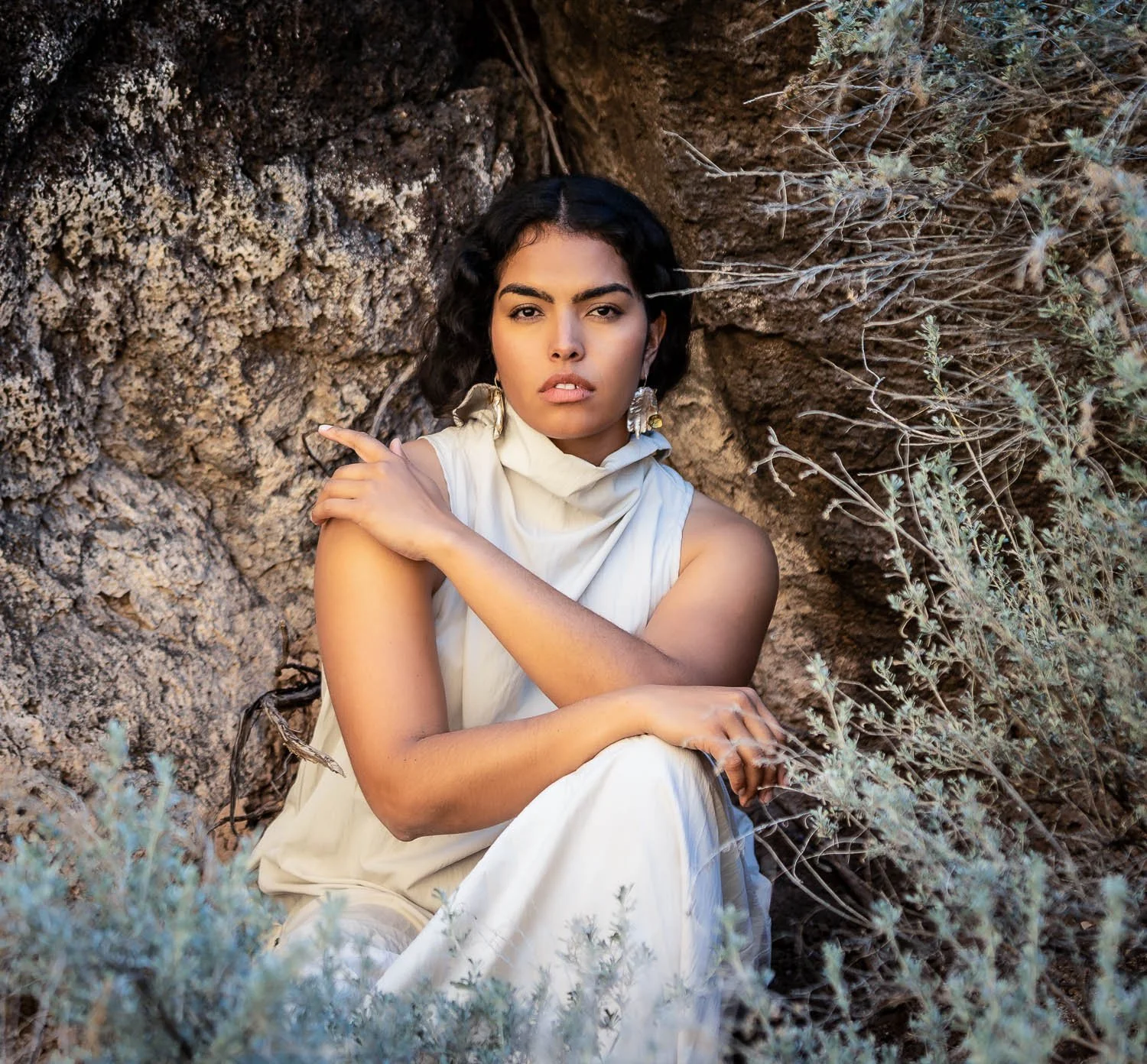 A woman with dark, curly hair and earrings sitting against a rocky, desert landscape with dry plants.
