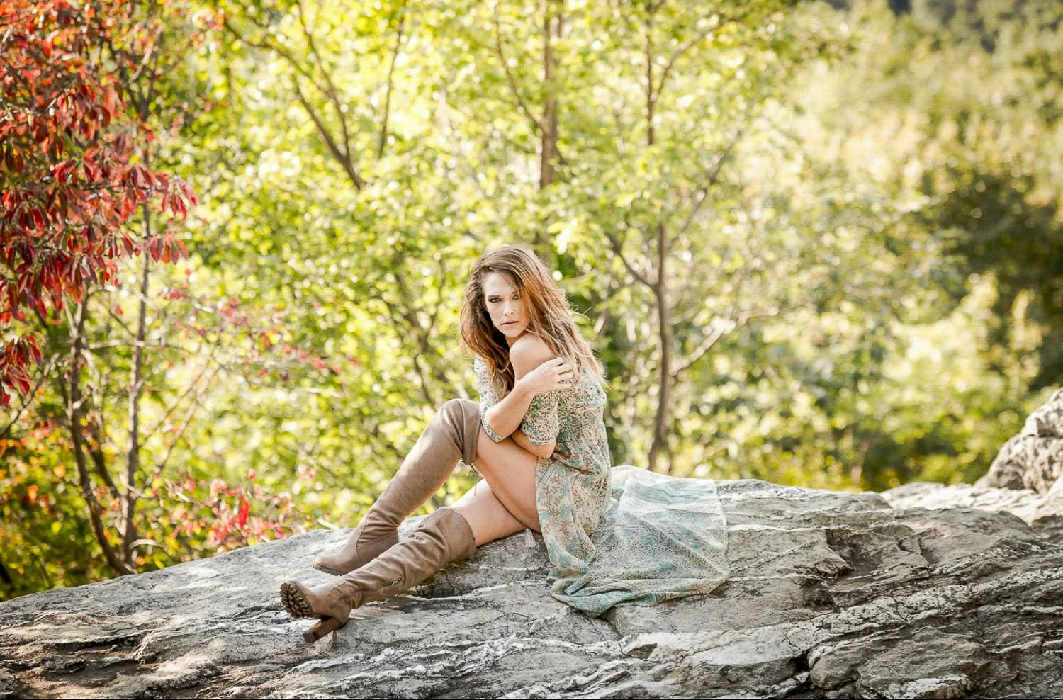 A woman sitting on a large rock outdoors with green trees and foliage in the background.