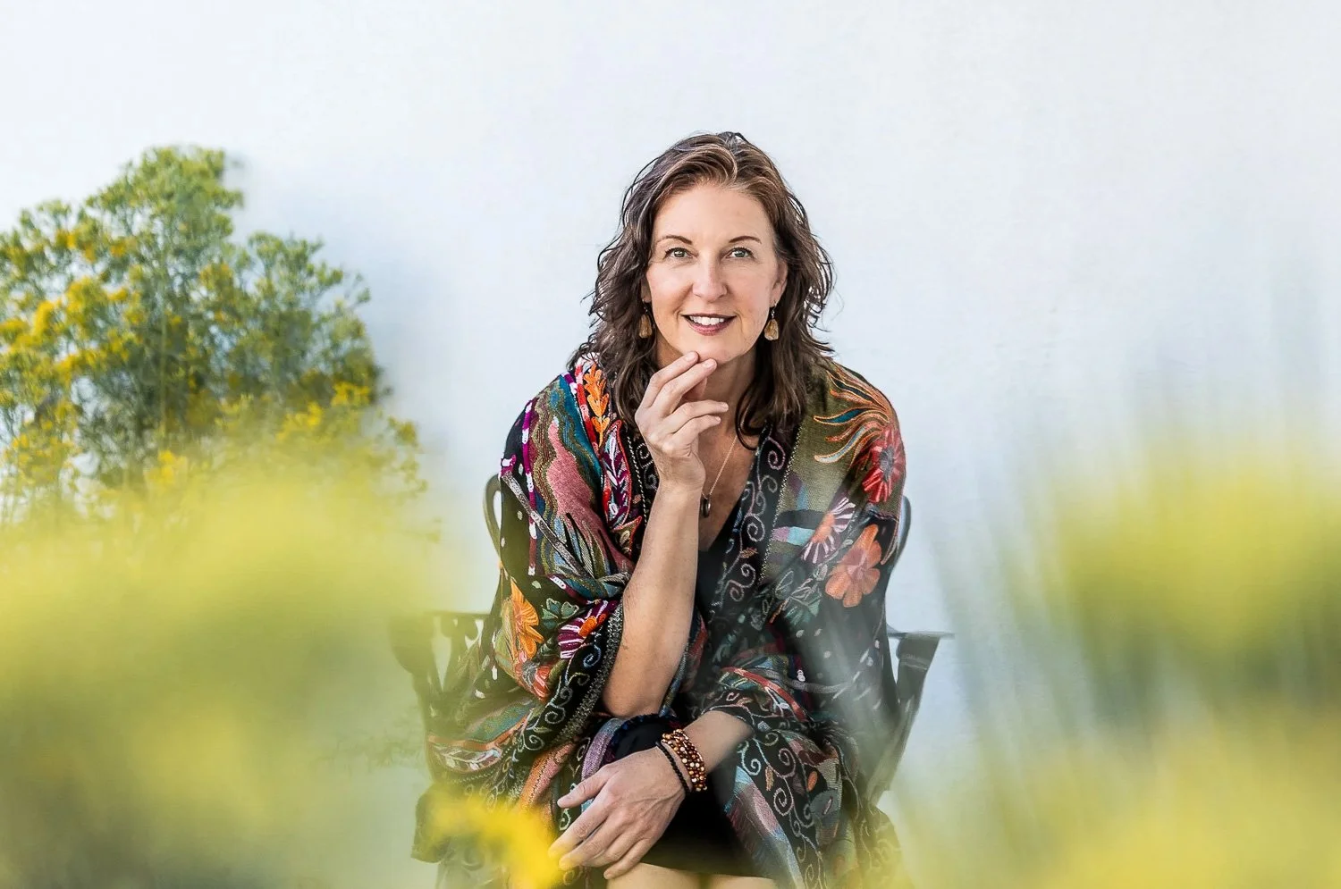 A woman with shoulder-length dark hair, wearing a colorful patterned shawl, sitting outdoors on a black chair, smiling and touching her face with her right hand, with yellow flowers in the foreground and a green tree and sky in the background.