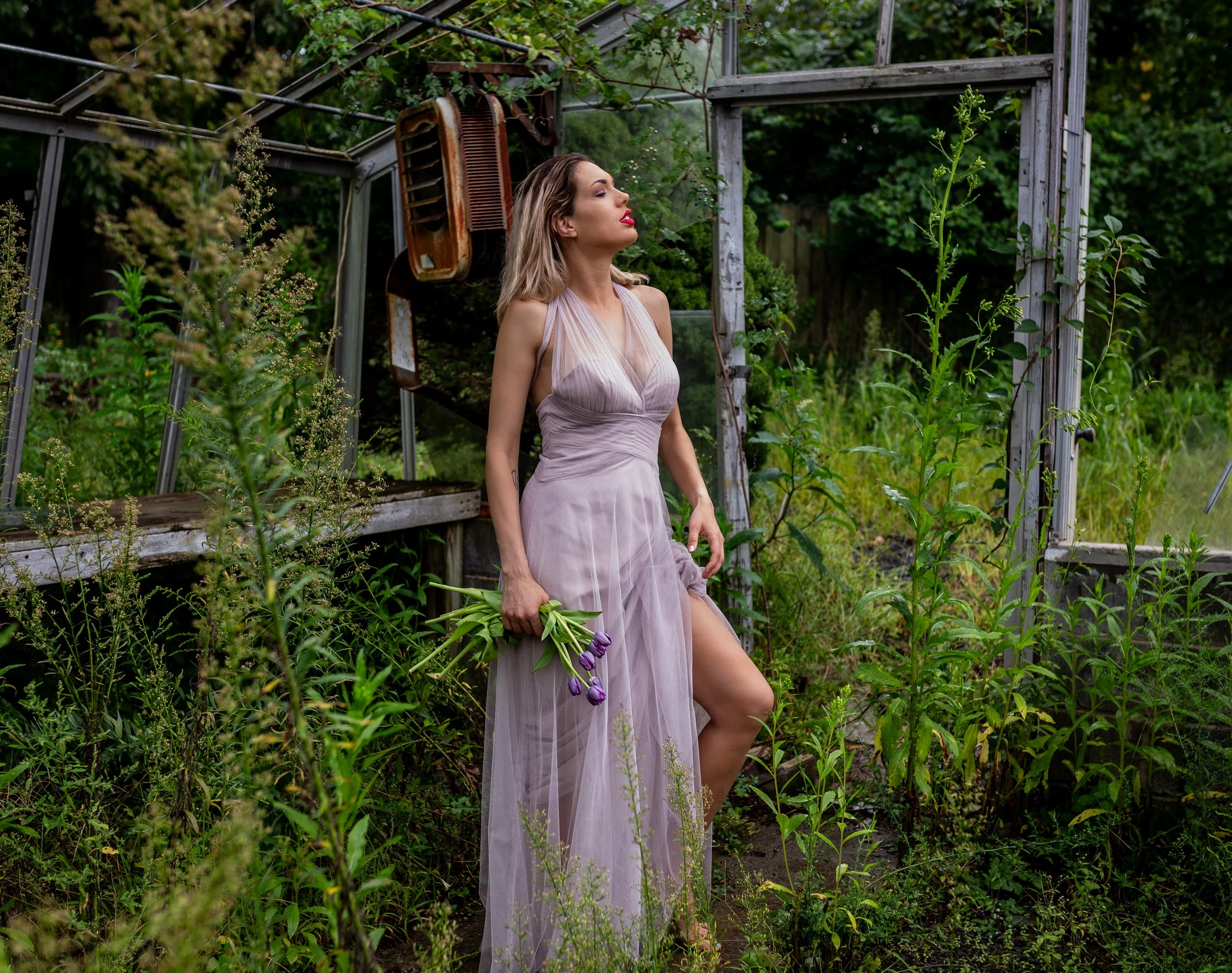 A woman in a flowy light purple dress holding purple tulips, standing in a lush green garden with overgrown plants and an old, weathered greenhouse in the background.