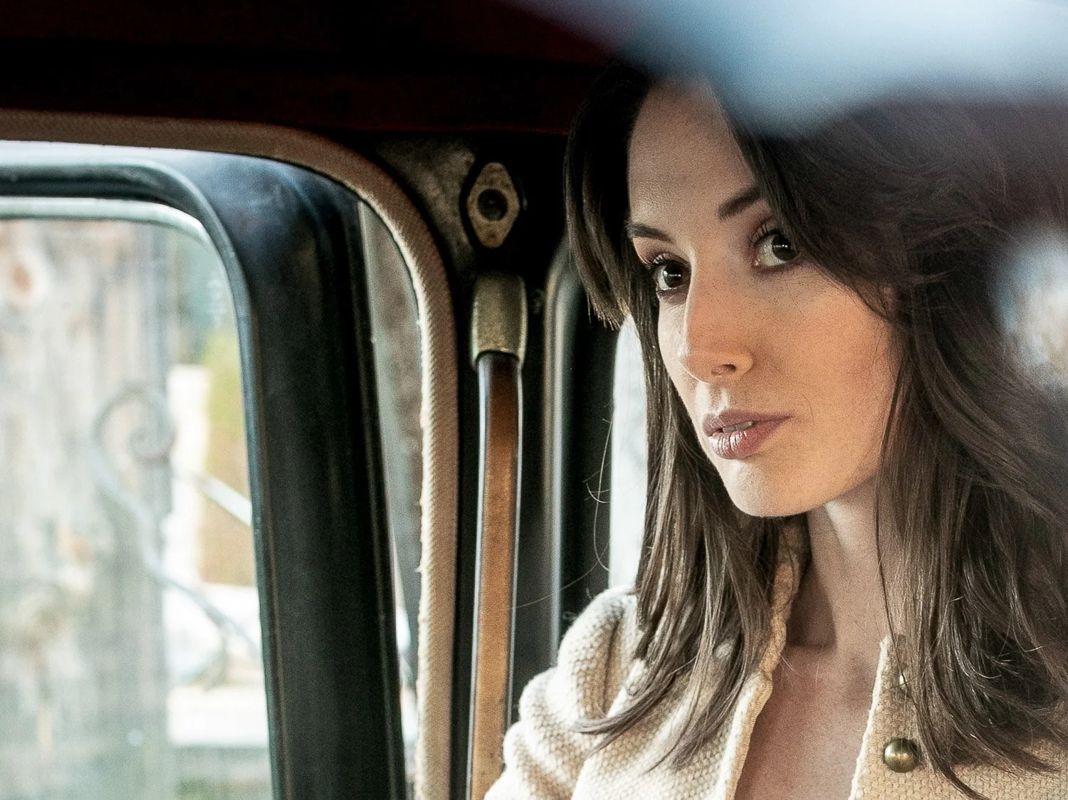 A woman with long brown hair and makeup sitting inside a vehicle, looking out the window.