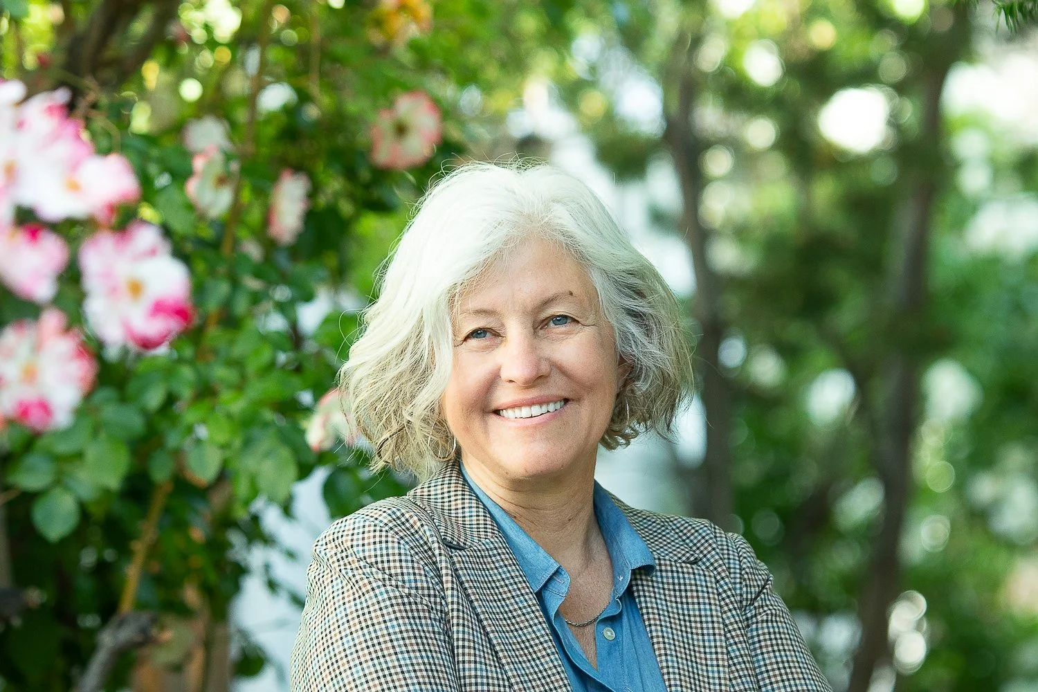A smiling woman with curly gray hair standing outdoors with greenery and pink flowers in the background.