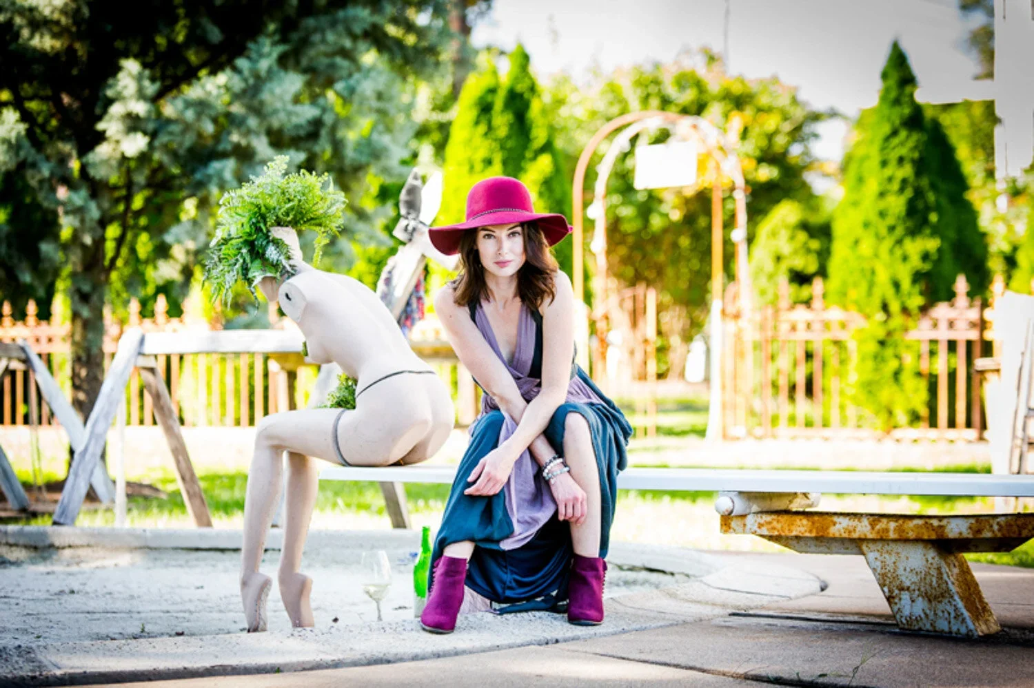 A woman sitting on a park bench wearing a large red hat, purple dress, and pink boots, with a white mannequin horse beside her in a park with lush green trees and a fence in the background.