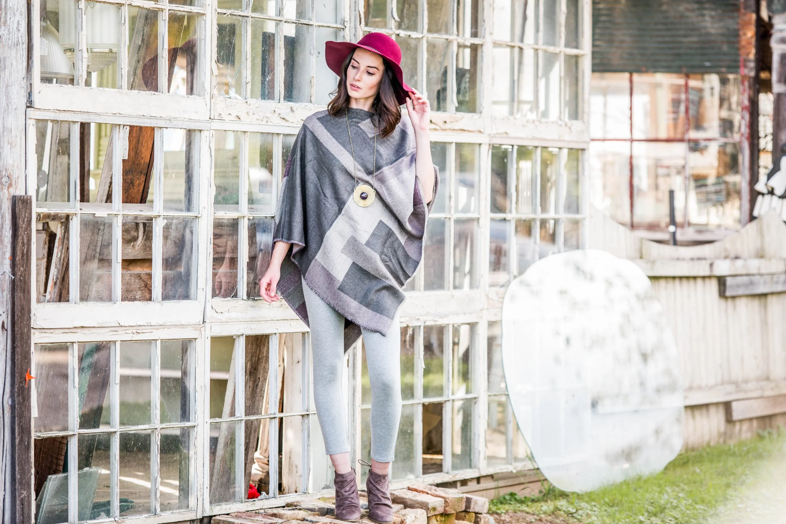 A woman wearing a pink wide-brimmed hat, gray patterned poncho, gray pants, and purple boots standing outside next to broken glass window panes in a rustic setting.