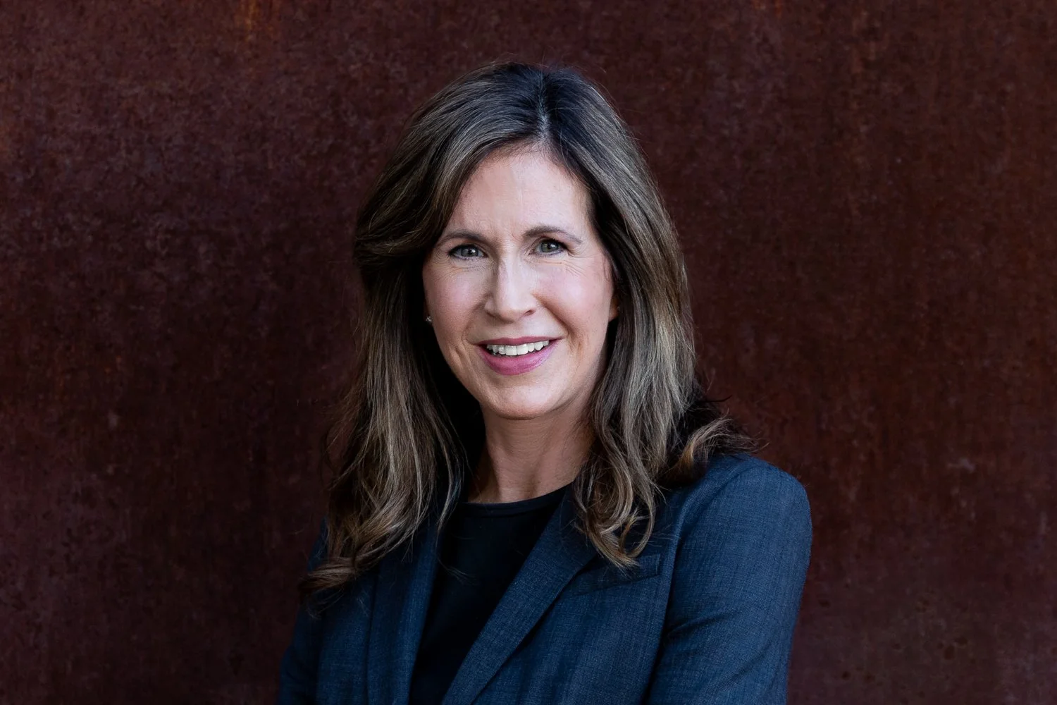 A woman with shoulder-length brown hair, smiling, wearing a dark blazer, standing against a textured dark red background.