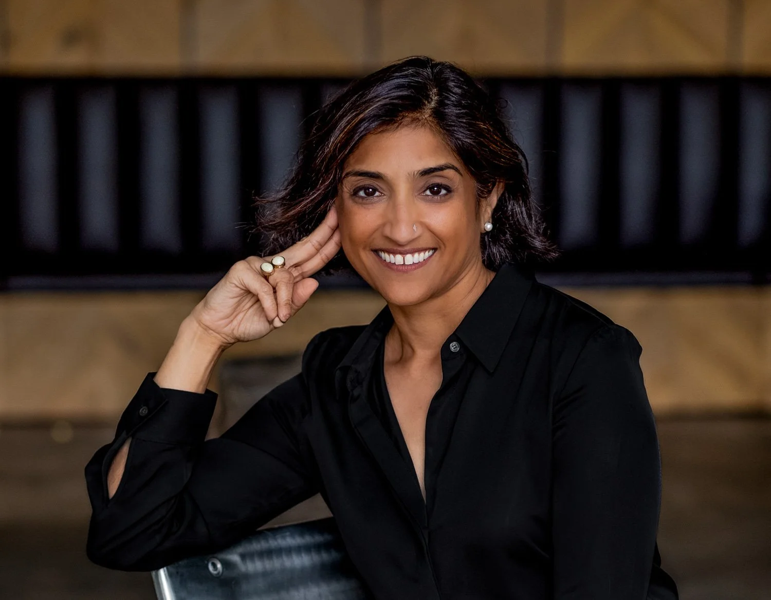 A woman with shoulder-length dark hair smiling and resting her face on her right hand, wearing a black button-up shirt and pearl earrings, sitting on a black chair in front of a wooden background with black vertical panels.