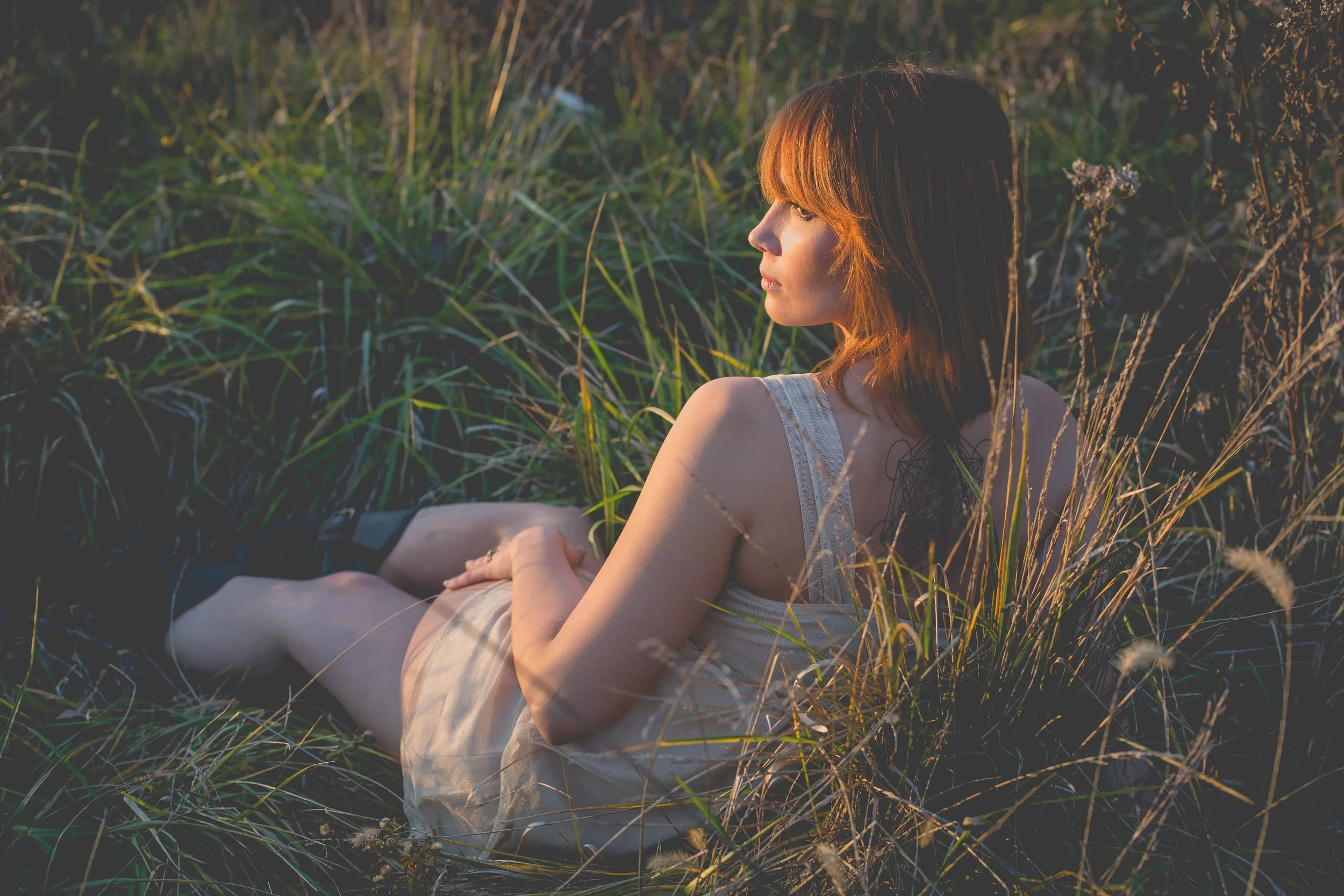 A young woman with red hair sitting in tall grass and wildflowers, looking to the side in sunset light.