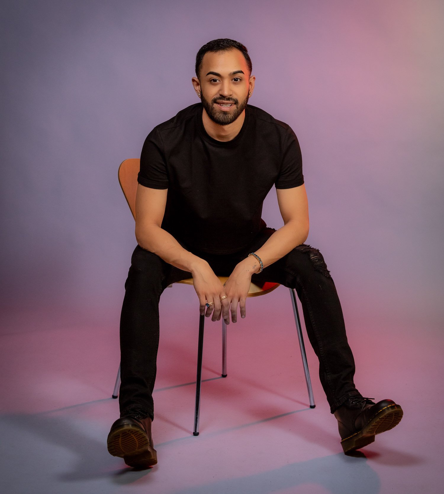 A man with dark hair and beard sitting on a chair against a pastel purple, pink, and blue background. He is wearing a black t-shirt, ripped black jeans, and black boots, smiling and looking at the camera.