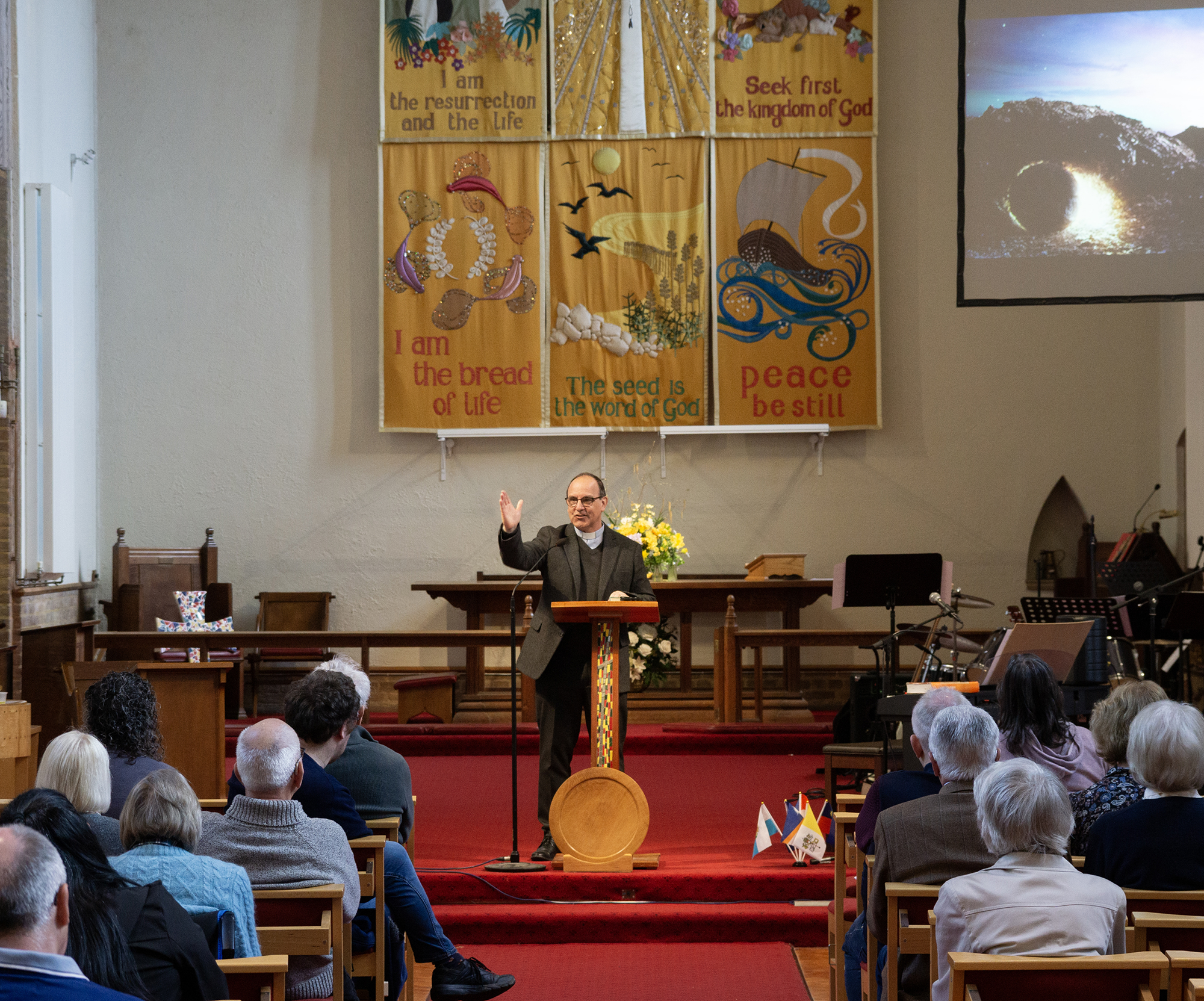 A Sunday service at Christchurch Orpington, vicar preaching sermon, church full of people, wall tapestry