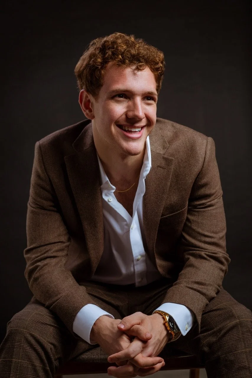 Smiling person in a brown suit seated against a dark background.