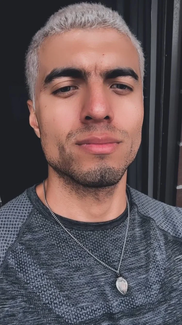 Close-up of a person with short silver hair, wearing a dark textured shirt and a silver necklace with a pendant.
