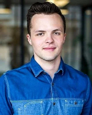 Man in blue denim shirt standing indoors with blurred background.