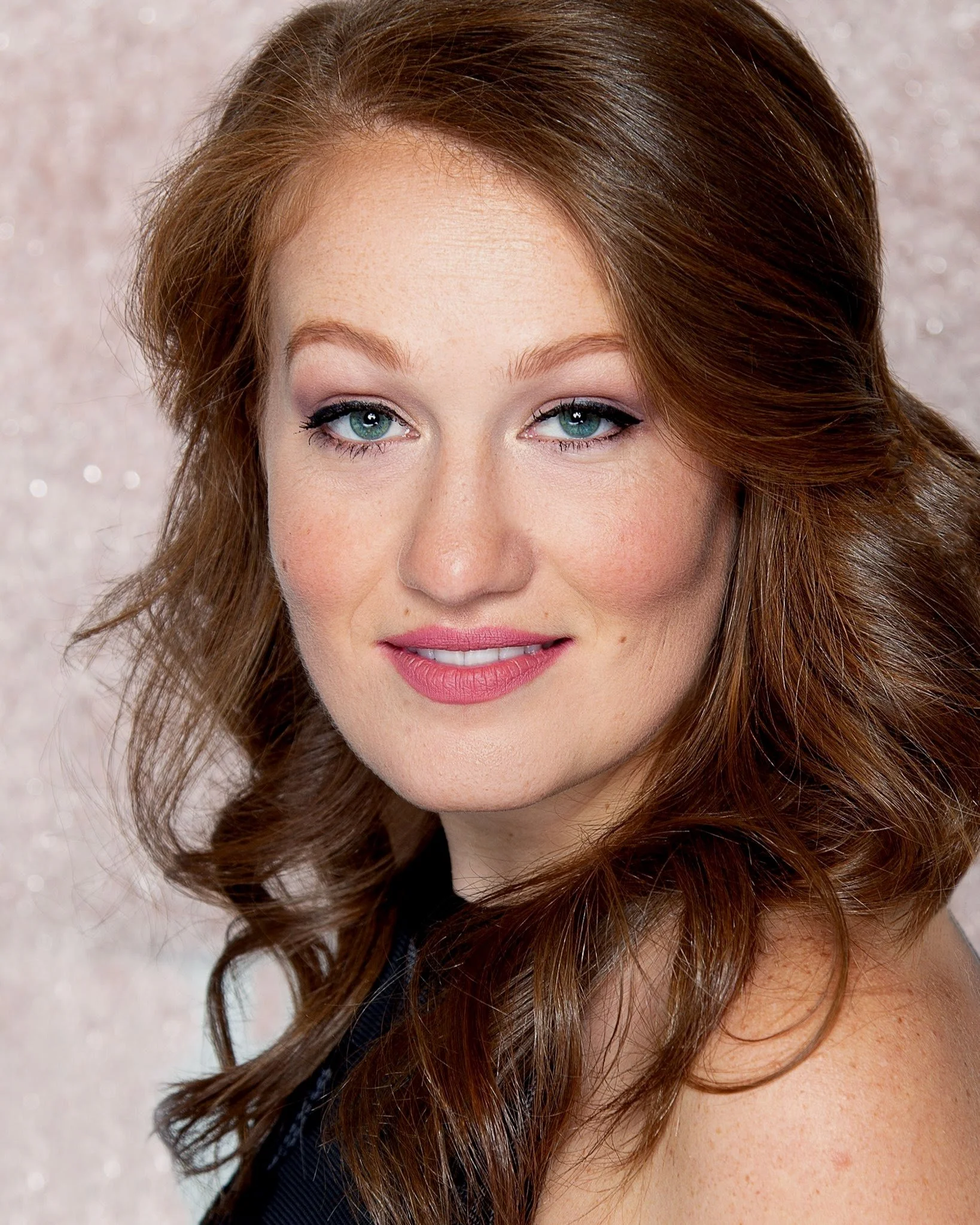 Portrait of a woman with red hair and a subtle smile, wearing makeup, against a beige sparkling background.