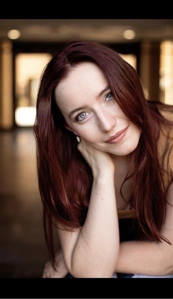 Portrait of a woman with long, reddish-brown hair, posing indoors. She is resting her chin on her hand and looking directly into the camera with a soft smile. The background is blurred, highlighting her face.